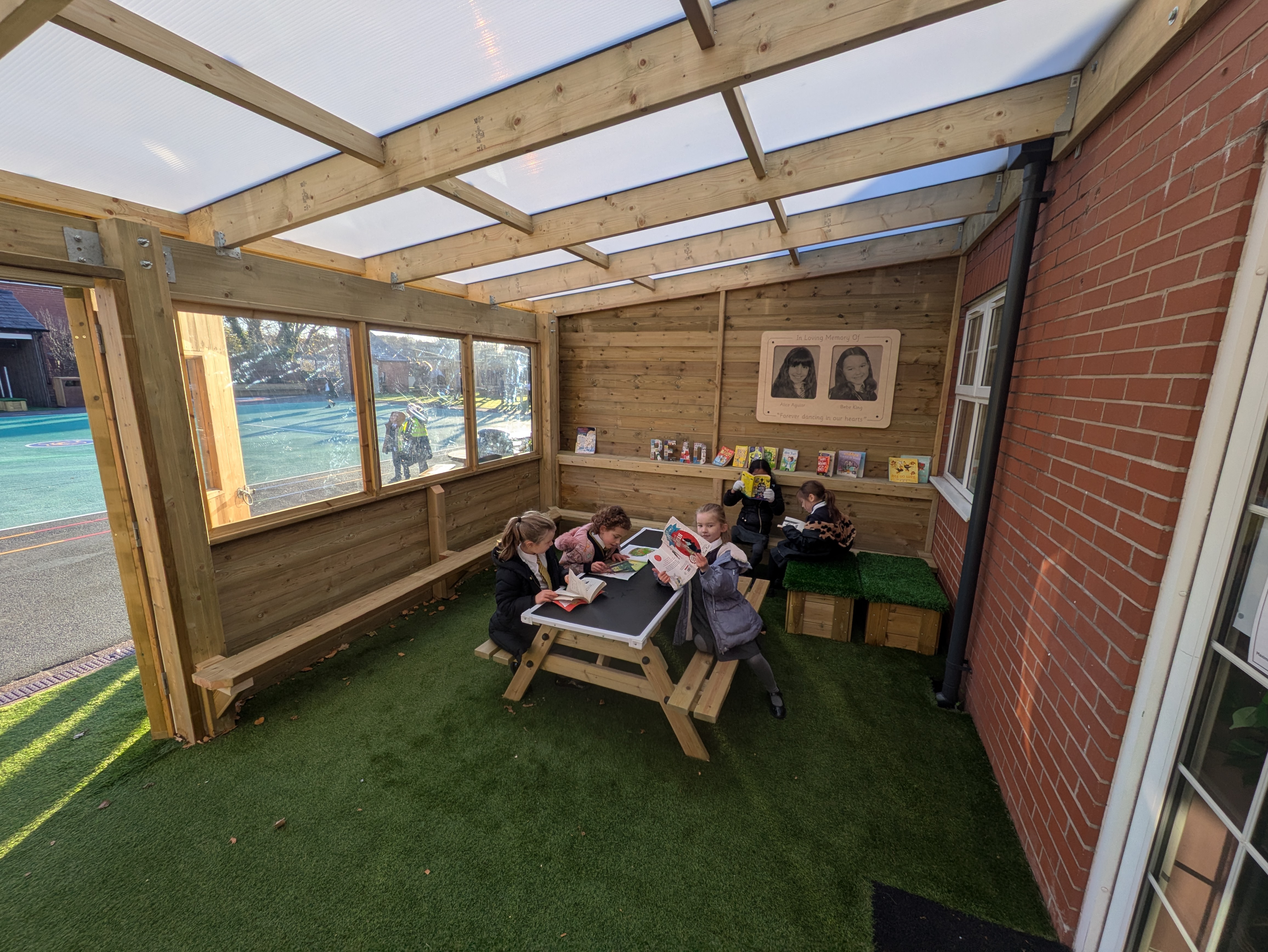 A group of children are sat under a canopy and are reading books.