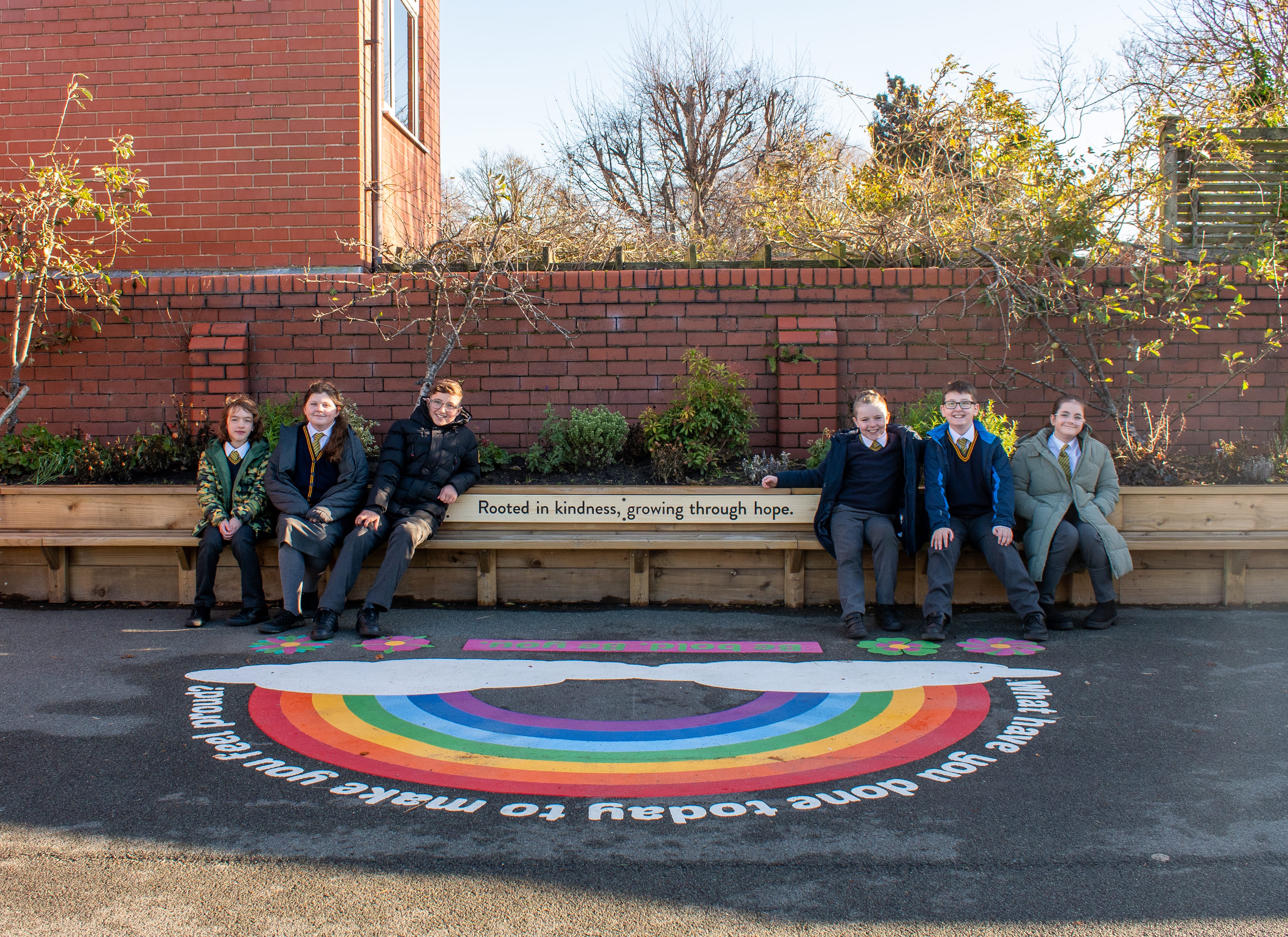 A group of children are sat on a bench, which has "Rooted in kindness, growing through hope." on it, with a thermoplastic rainbow in front of it.