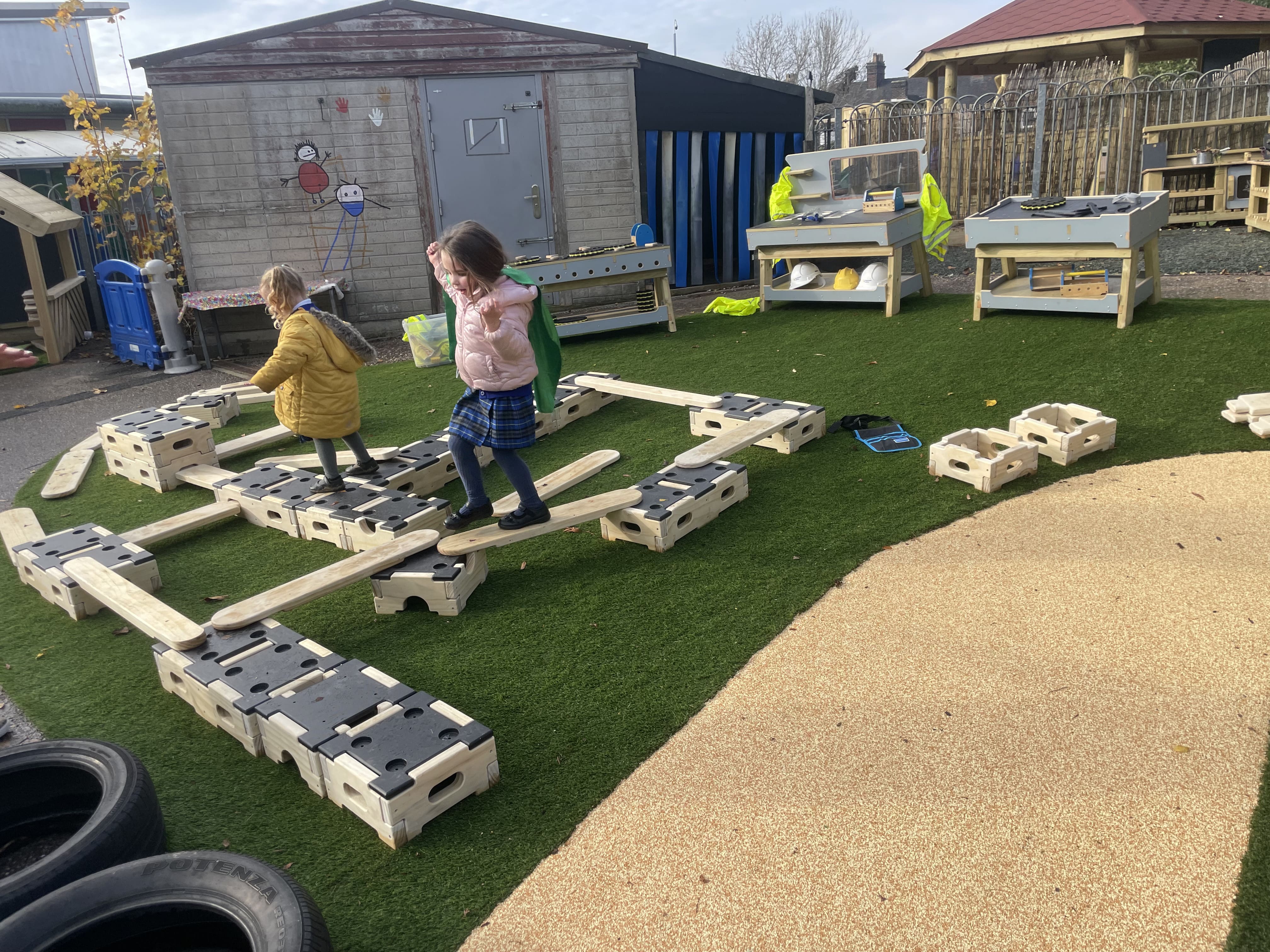 A few children are balancing across a Play Builder Engineer Set, that has been placed on top of artificial grass at Kingsland CofE.
