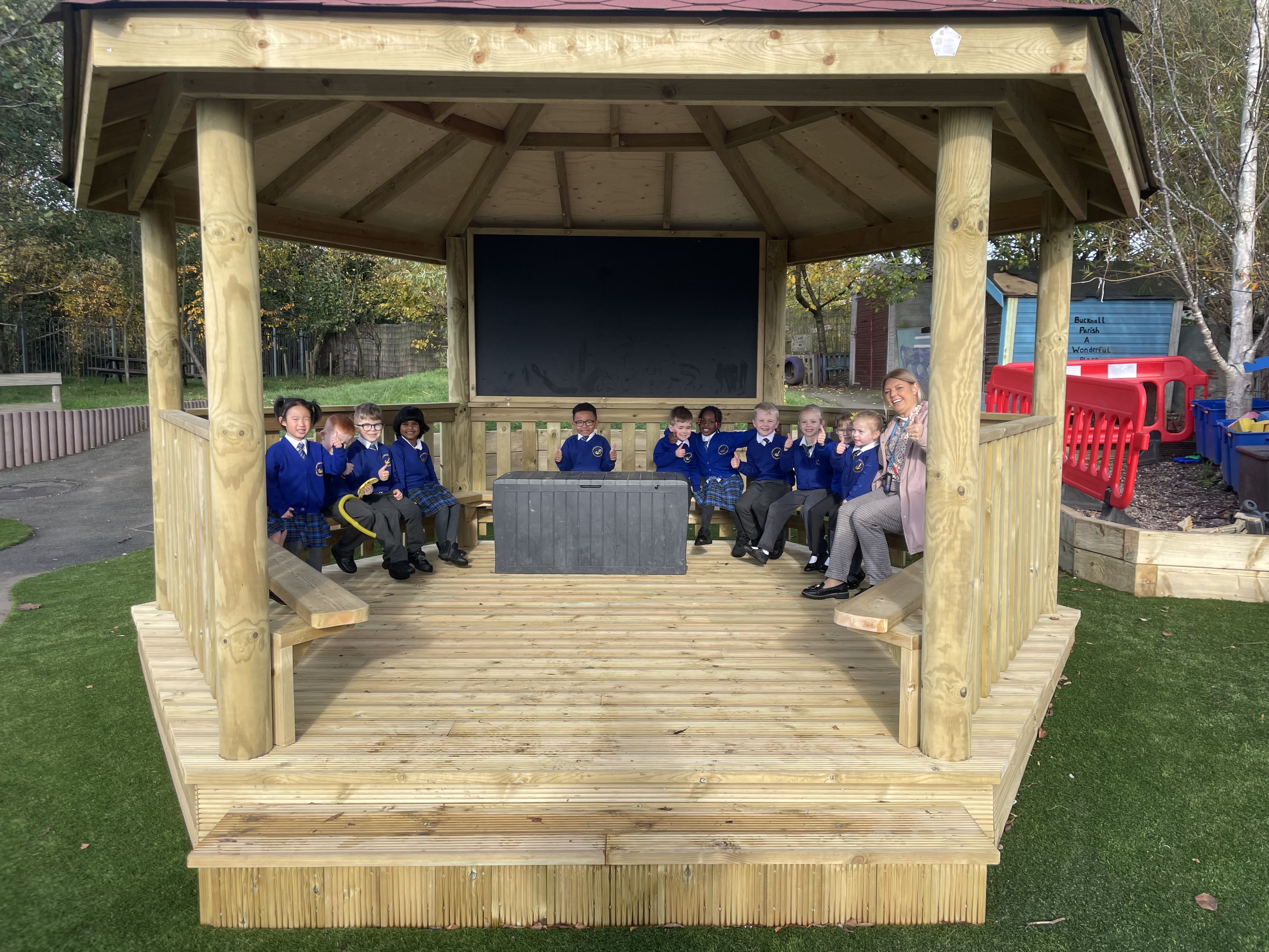 A group photo of the children sat inside a gazebo, with the teacher, all smiling at the camera and giving a thumbsup!