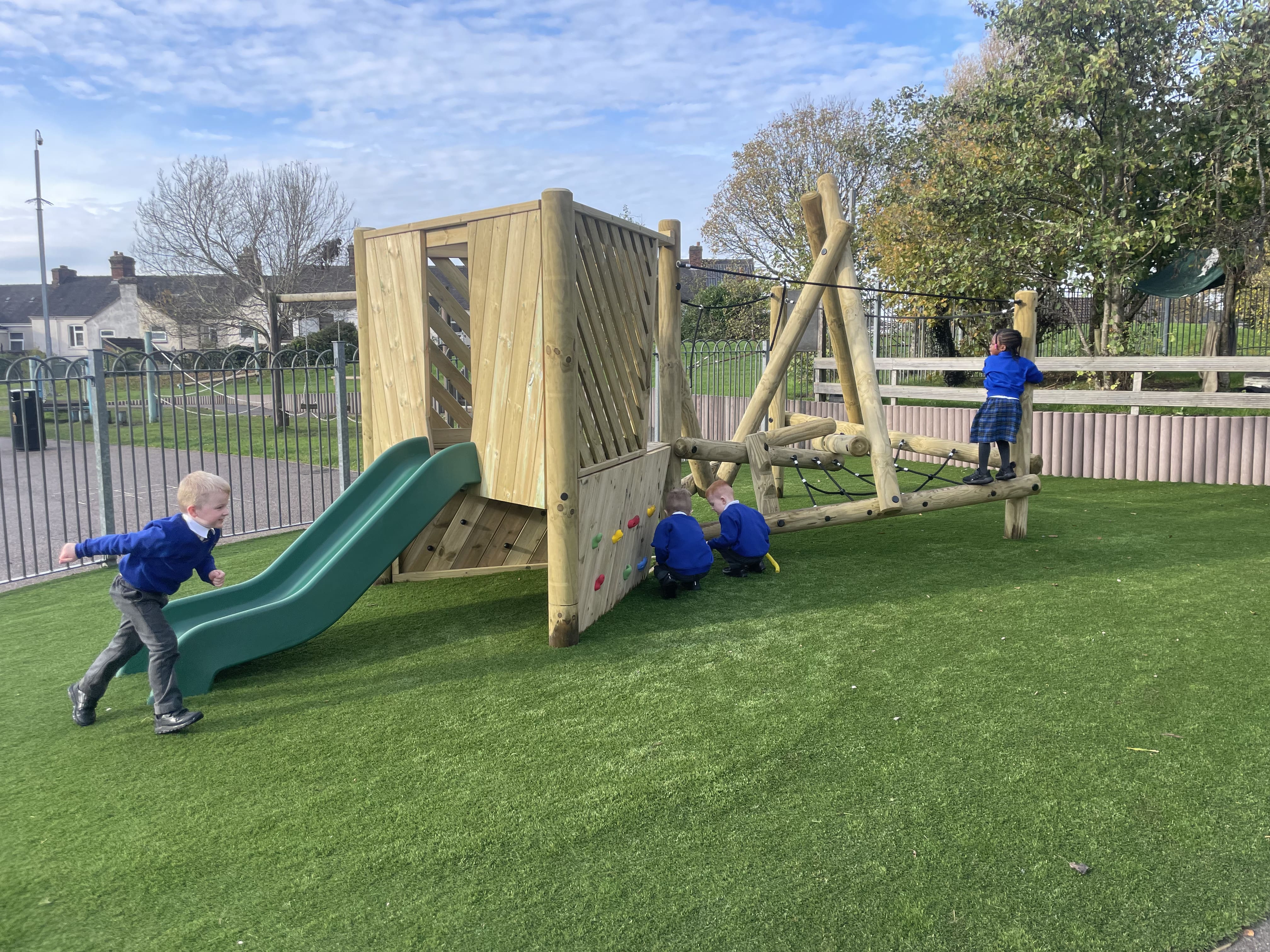 A climbing frame with an attached platform and slide, with a few children playing with the equipment on artificial grass.
