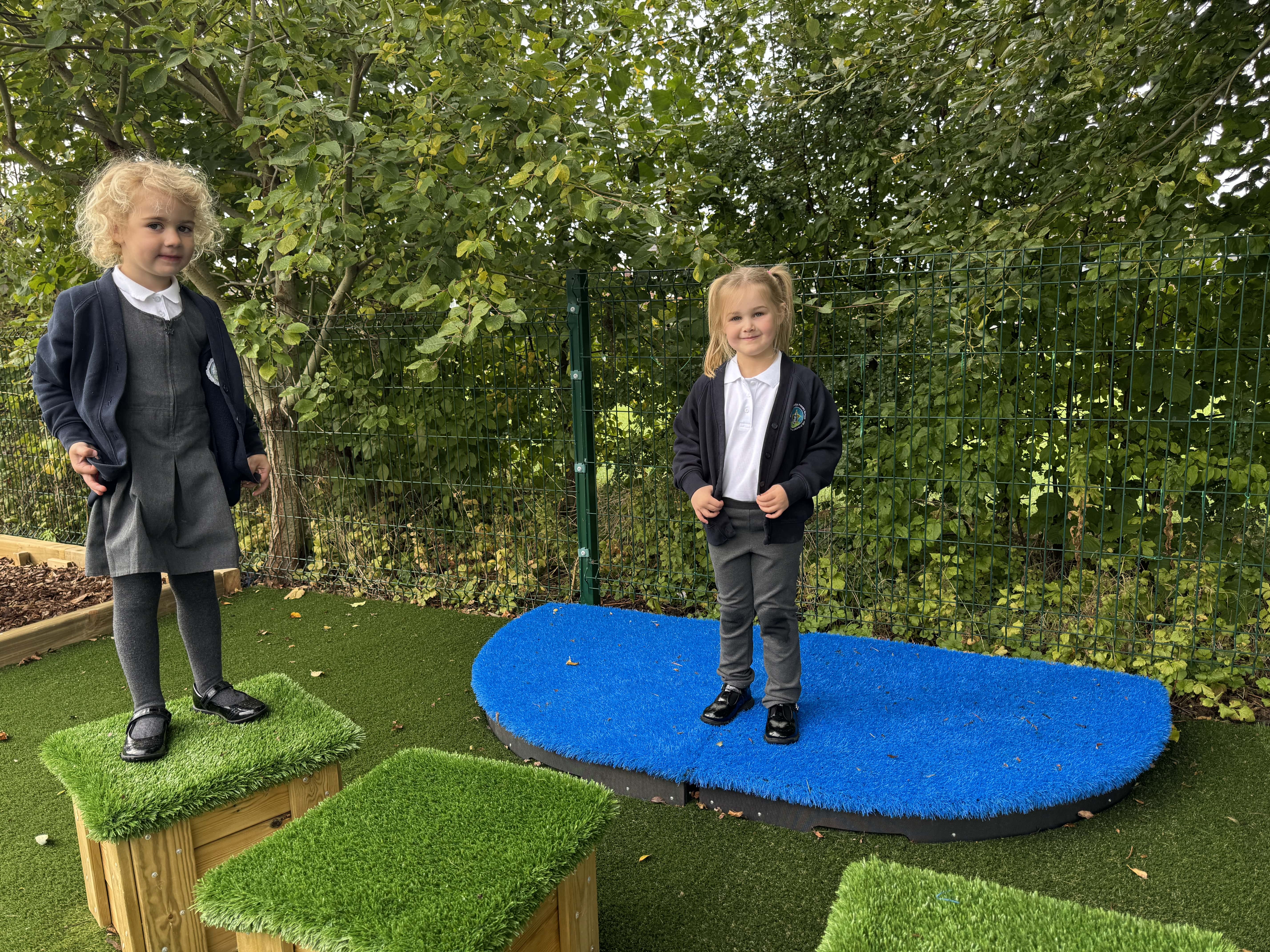 A girl is stood on top of an artificial grass seat, as another girl is stood on a dual freestanding blue stage, both smiling at the camera.