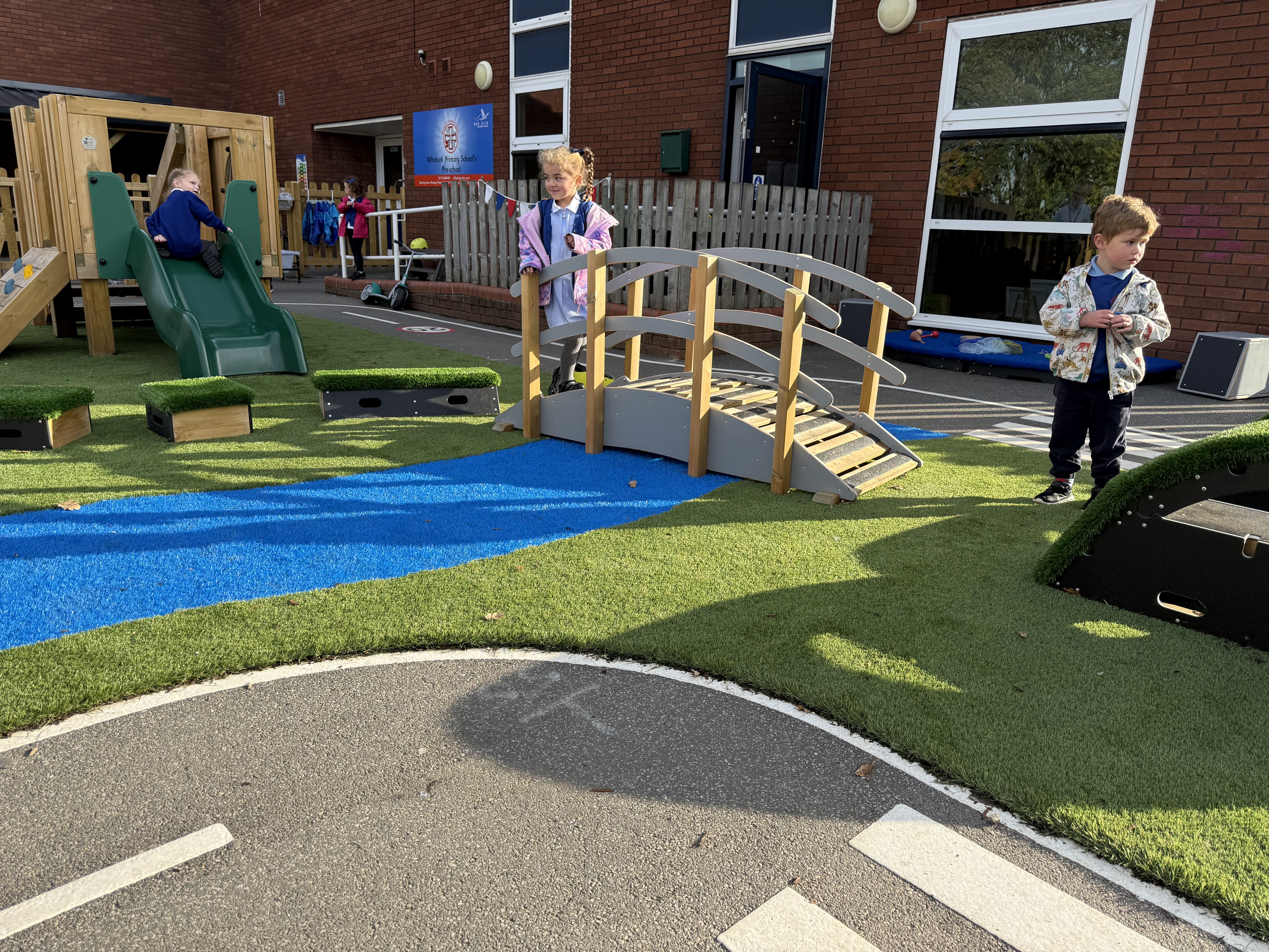 A group of children are walking on artificial grass, as a little girl stands on top of the freestanding play bridge.