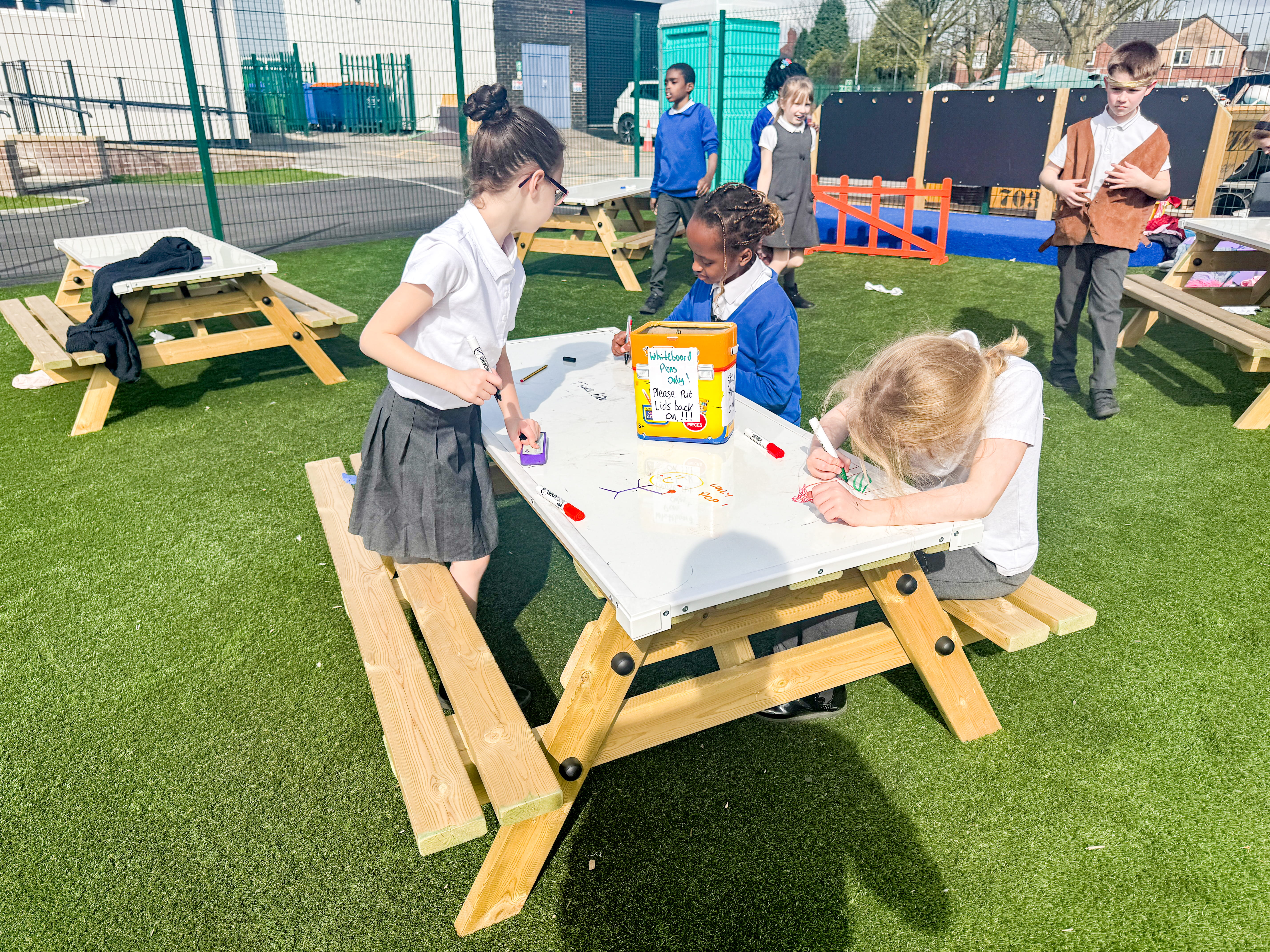 Three girls are sat at a Picnic Bench with a Whiteboard top, drawing on it and talking with each other.