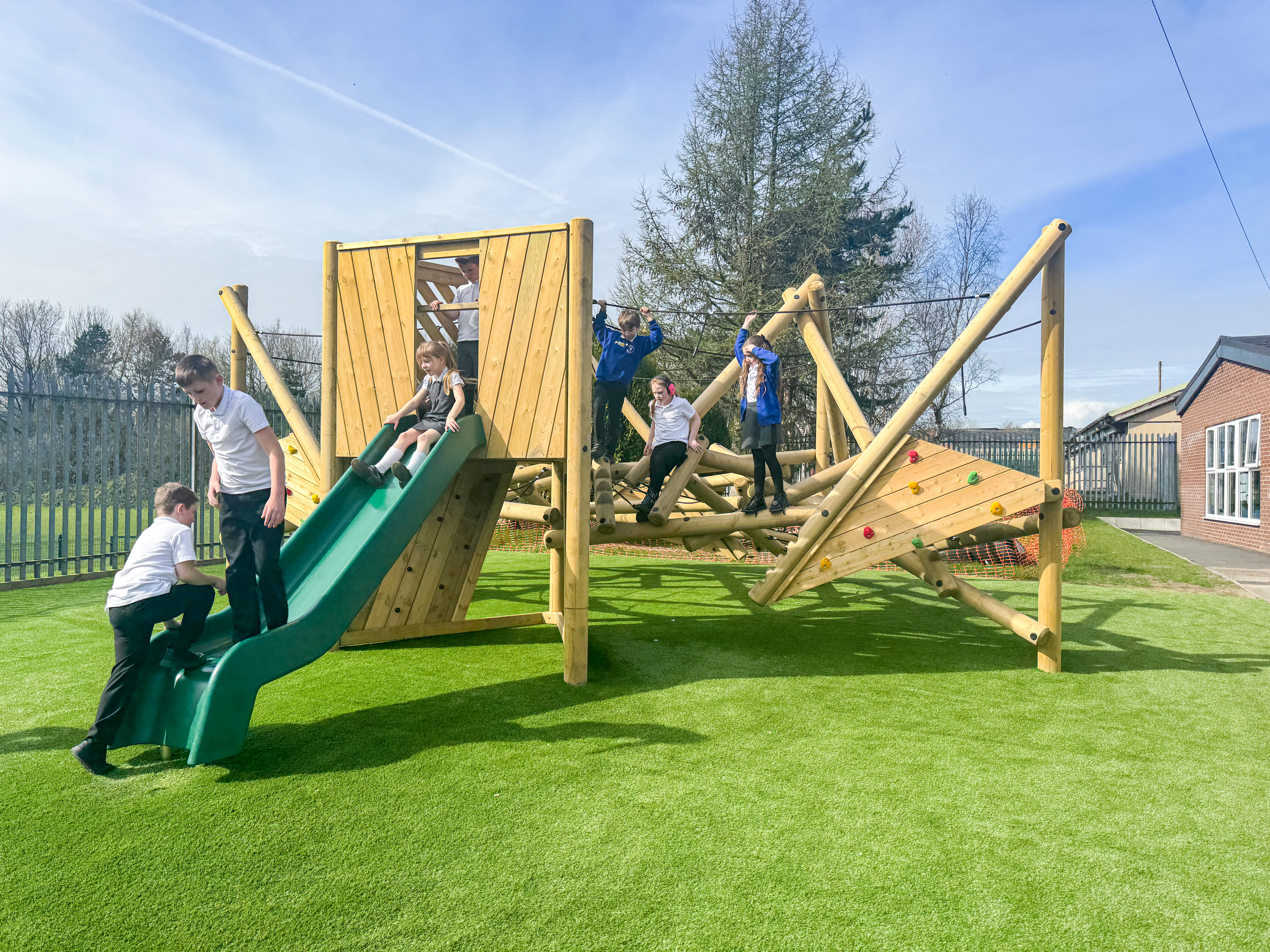 A large group of children are exploring the Crinkle Crags climbing frame, with a few children standing on the platform and sliding down the slide.