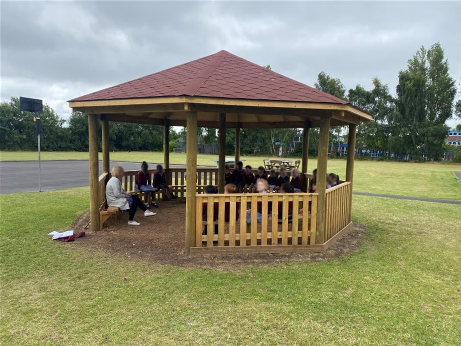 children sit on the benches around the inside of the outdoor classroom and listen to the teacher who stands at the entrance wearing a coat and talking to them