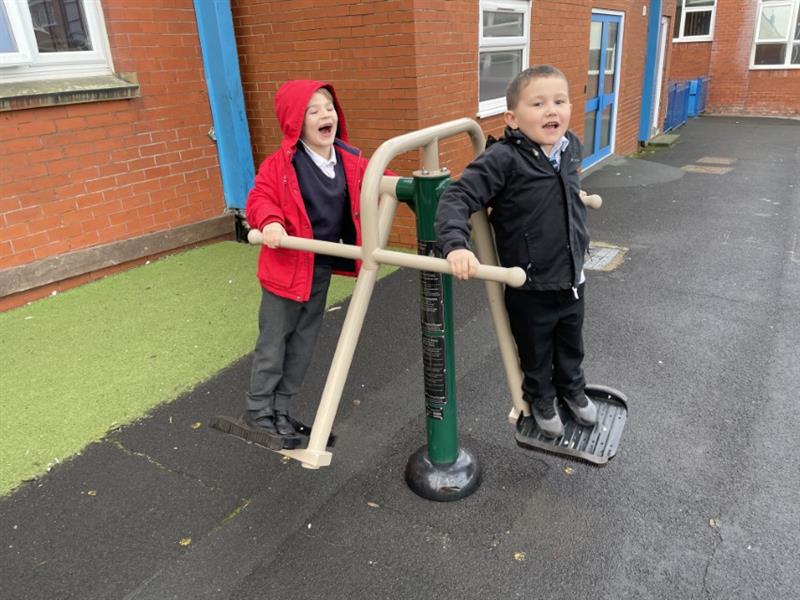 Two KS1 boys, one in a red coat with his hood up and one in a black coat, using a Double Air Walker and laughing in the playground