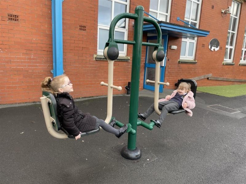 Two KS2 children in coats using a seated leg press in their school playground