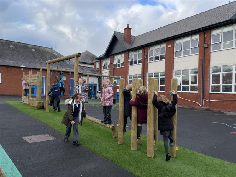 Nine children wearing coats moving across the nine challenges featured on their playground trim trail installed with artificial grass underneath