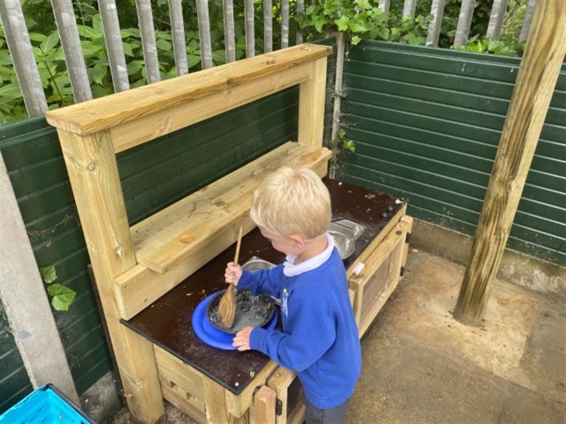 A reception child in a blue school jumper mixing mud on the mini mud kitchen using a wooden spoon