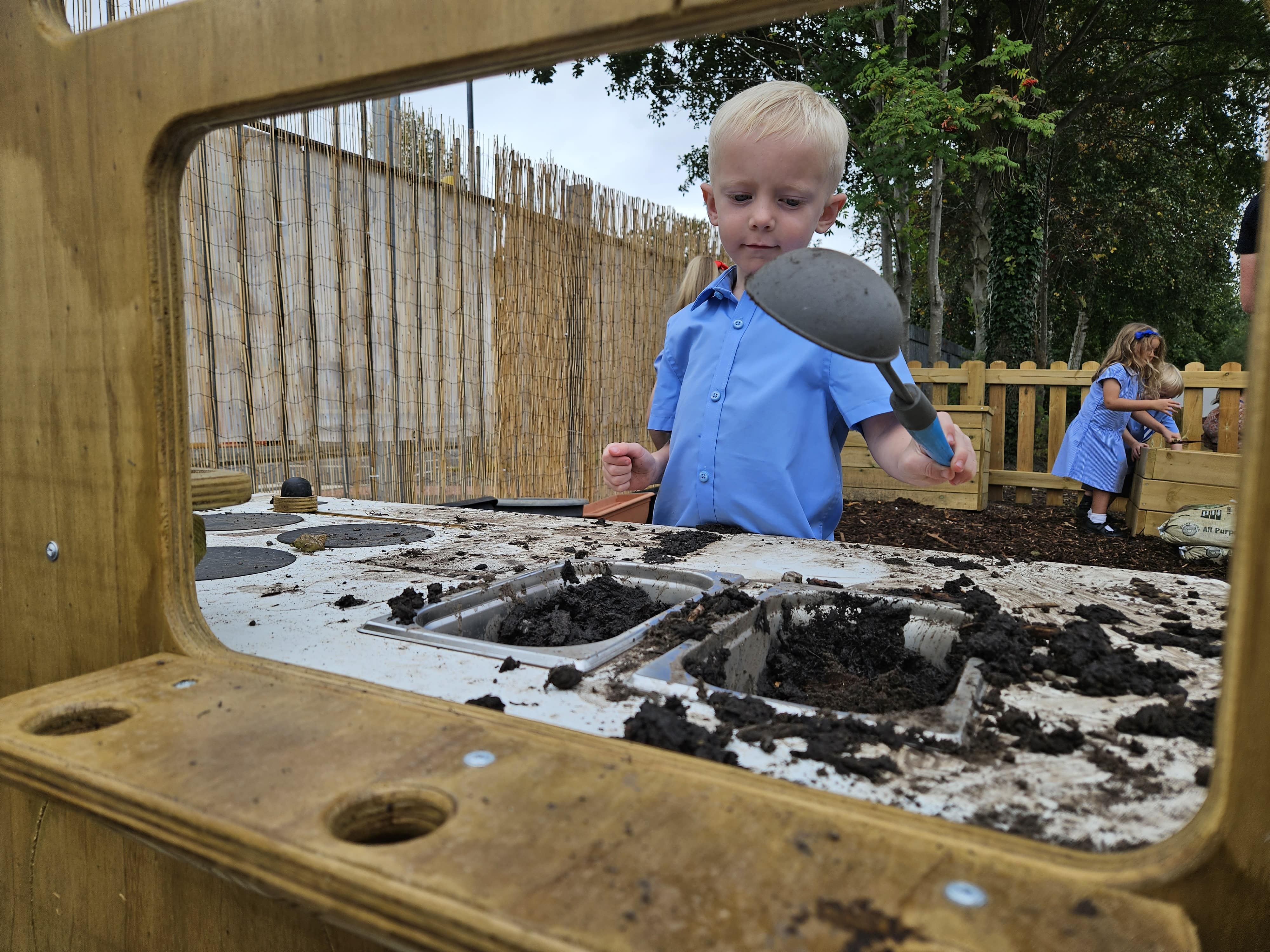 A young child playing with a mud kitchen. The young boy is using a soup spoon as they scoop mud in and out of the containers,