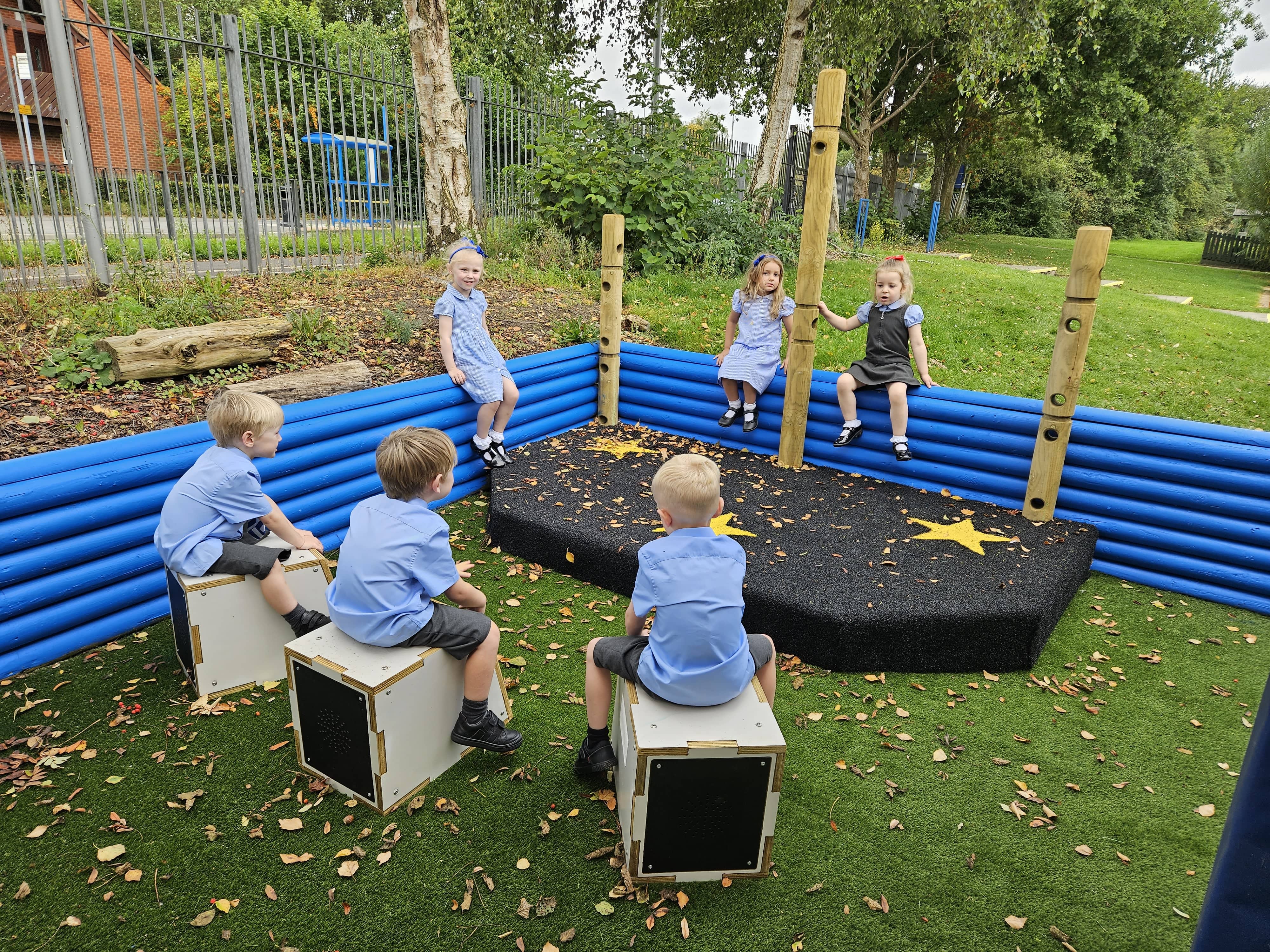 6 children are playing with the outdoor Performance Stage with Den Posts. The Performance Stage is black with star patterns on it and 3 children are sat on the fence that is behind it.