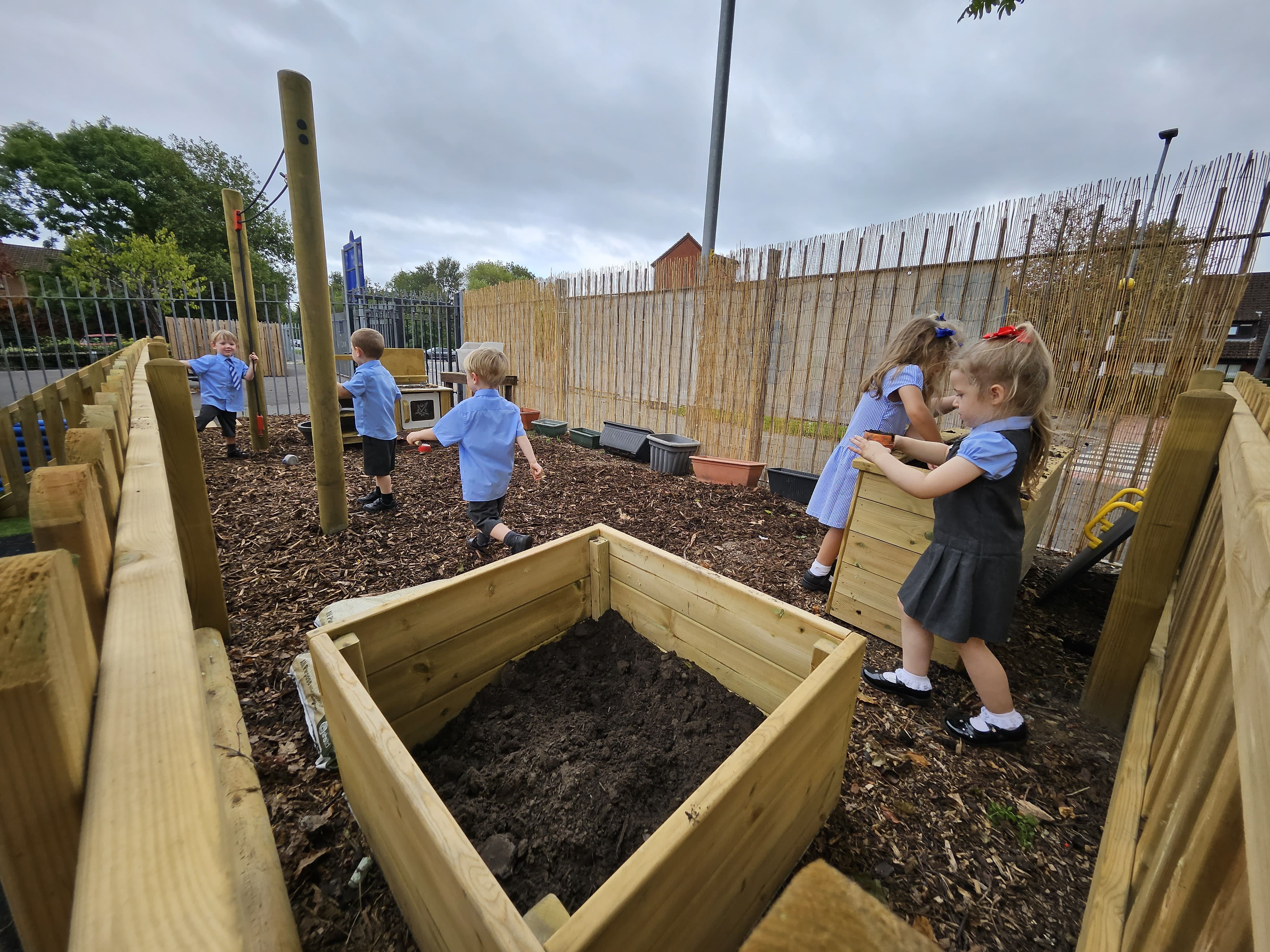 5 Children are walking around the messy area which has been built by Pentagon Play. 2 Planters are full of mud and there's a pulley and mud kitchen on the other side of the area. The children are playing with all the equipment as they engage in messy play.