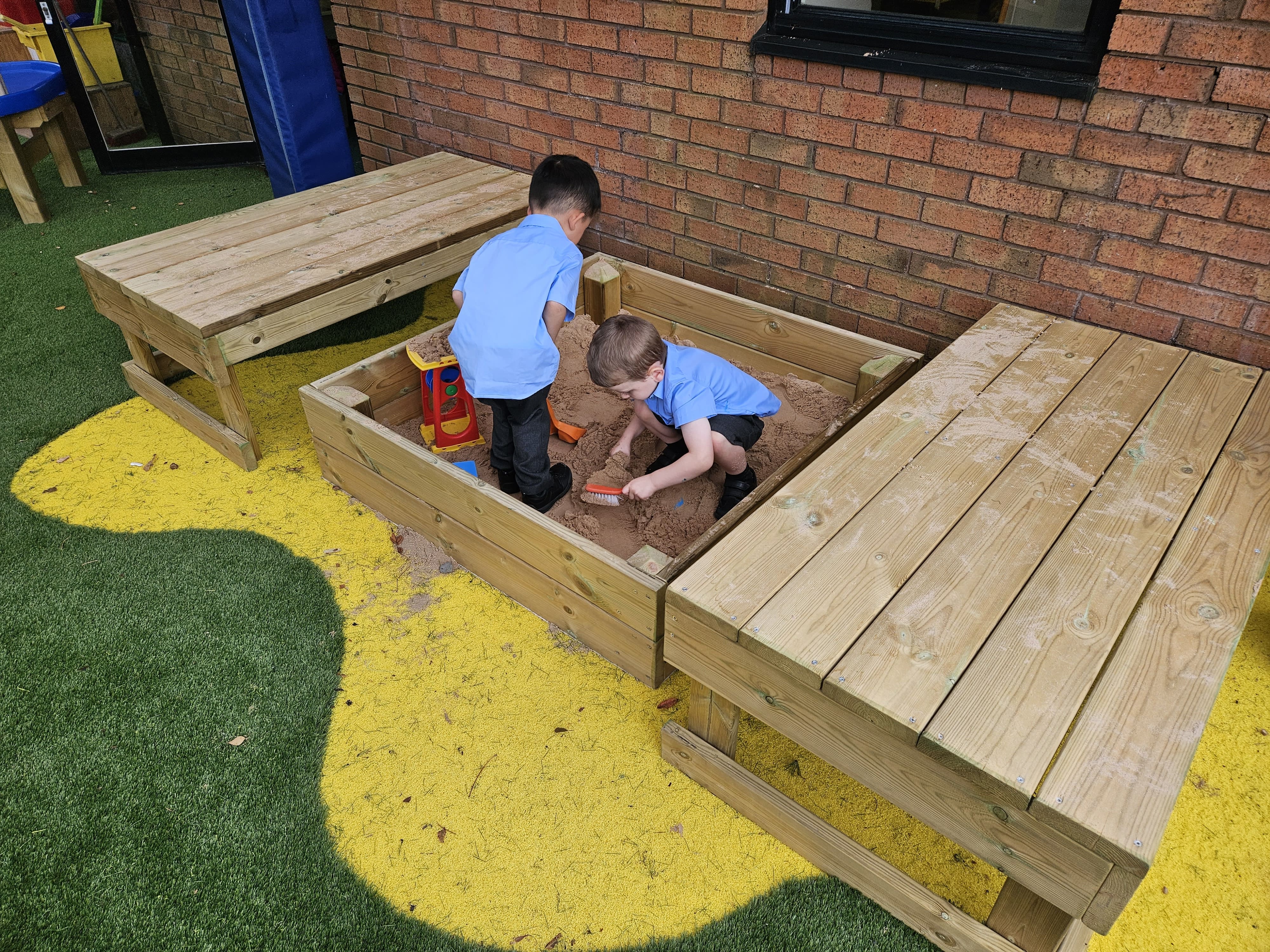 2 children are playing with the Pentagon sandbox. The kids are using some different tools, like a brush and shovel, to move the sand around. Outside the box, the artificial grass can be seen, creating a colourful environment,