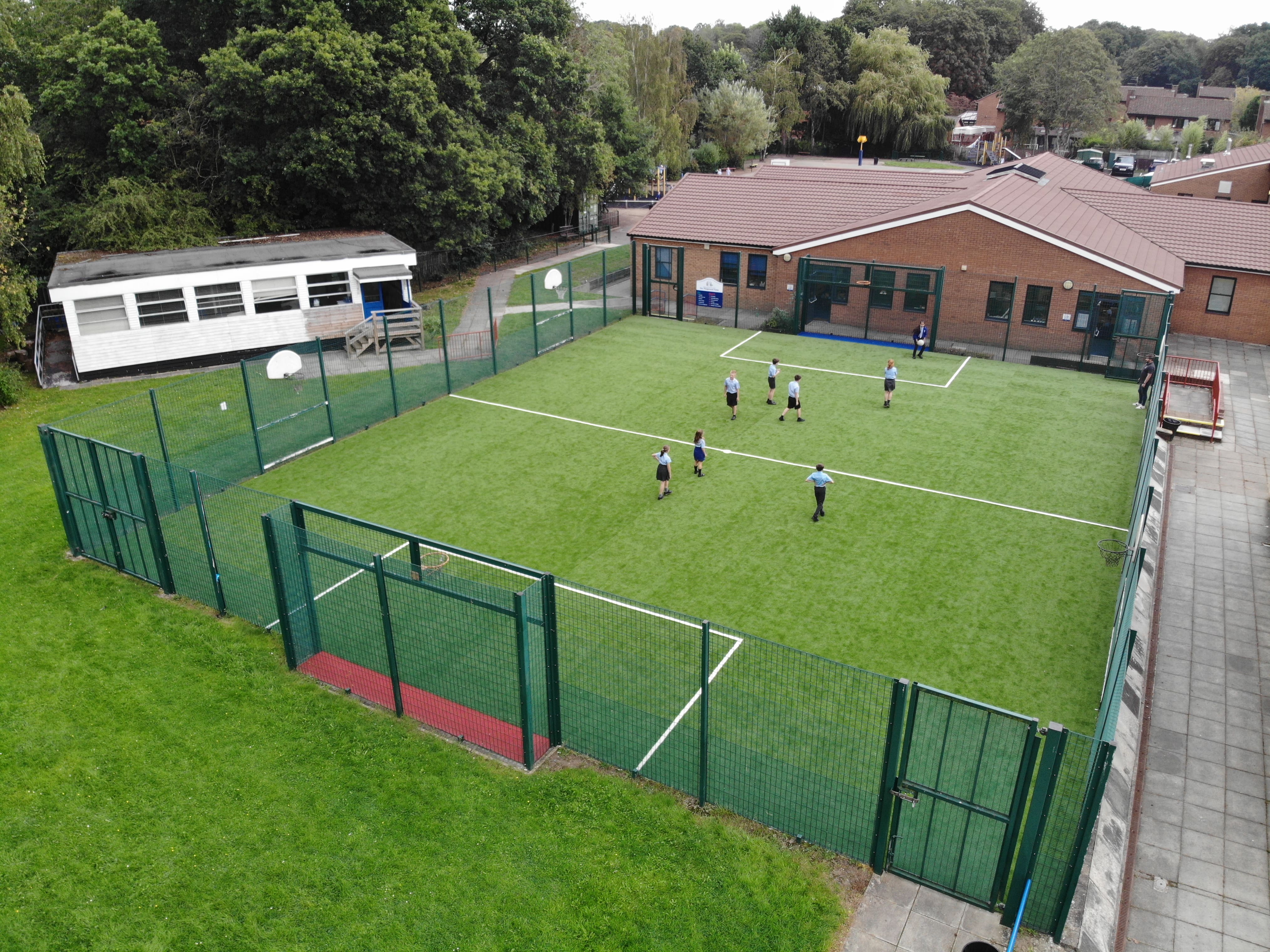 A drone shot of the MUGA from above. The MUGA is green with white lines which are football markings. The floor of both goals are different colours, that being red and blue. There are basketball hoops on the green fence that goes around the pitch.