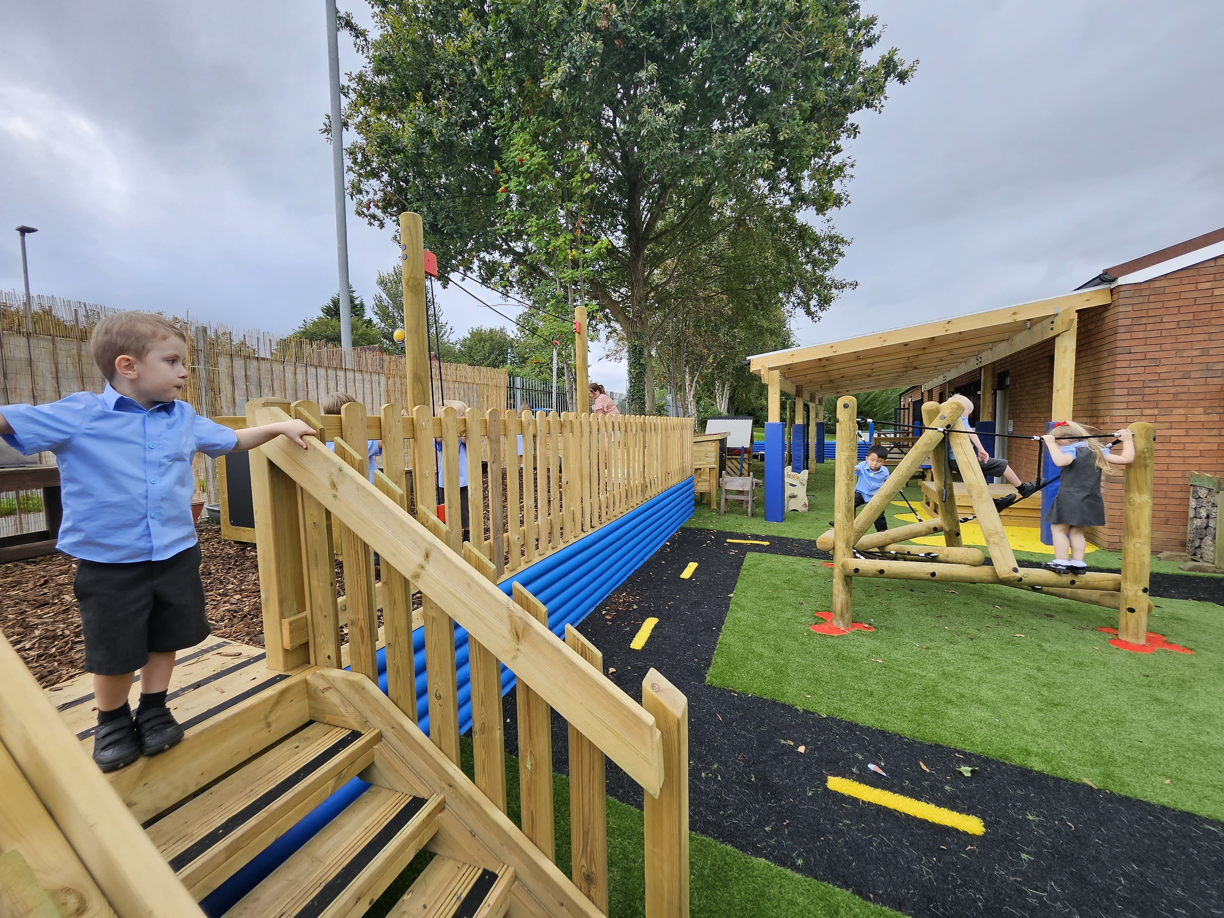 A child is stood on timber states as he looks over to the active play area. At the top of the staircase, where the child is stood, is where the messy play area is. The active play area has the playturf deluxe artificial grass flooring whilst the messy play area has the wooden bark chops as it's flooring.