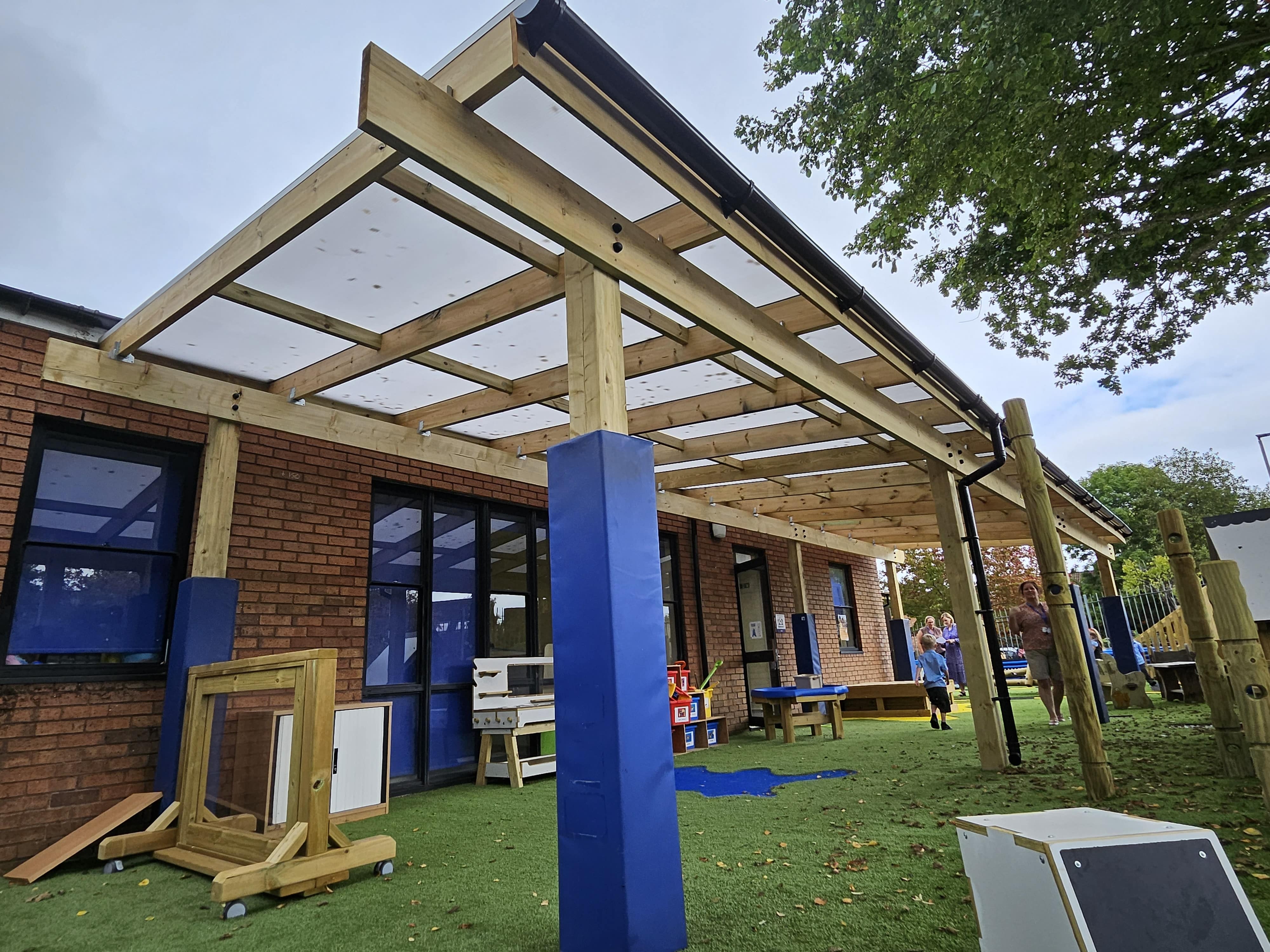 A freestanding timber canopy attached to the school. Underneath the canopy is a wide variety of different play equipment, designed to help children develop their skills. The flooring is Playturf Artificial Grass.