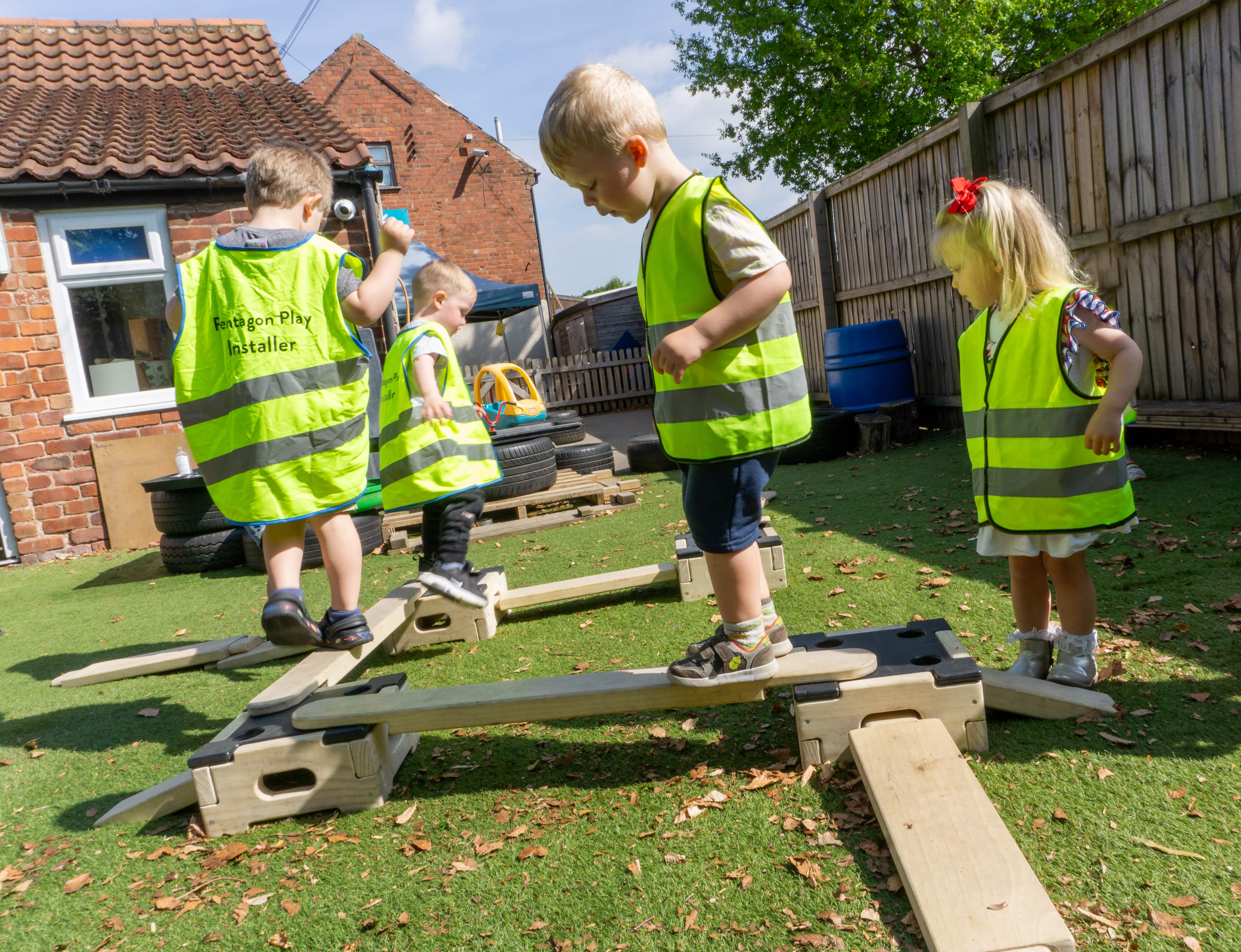 4 children are wearing hi-vis jackets which say "Pentagon Play Installer" on the back of them. The children have built a small obstacle course with the Play Builder set, which is set up in the playground of Kindred White Post.