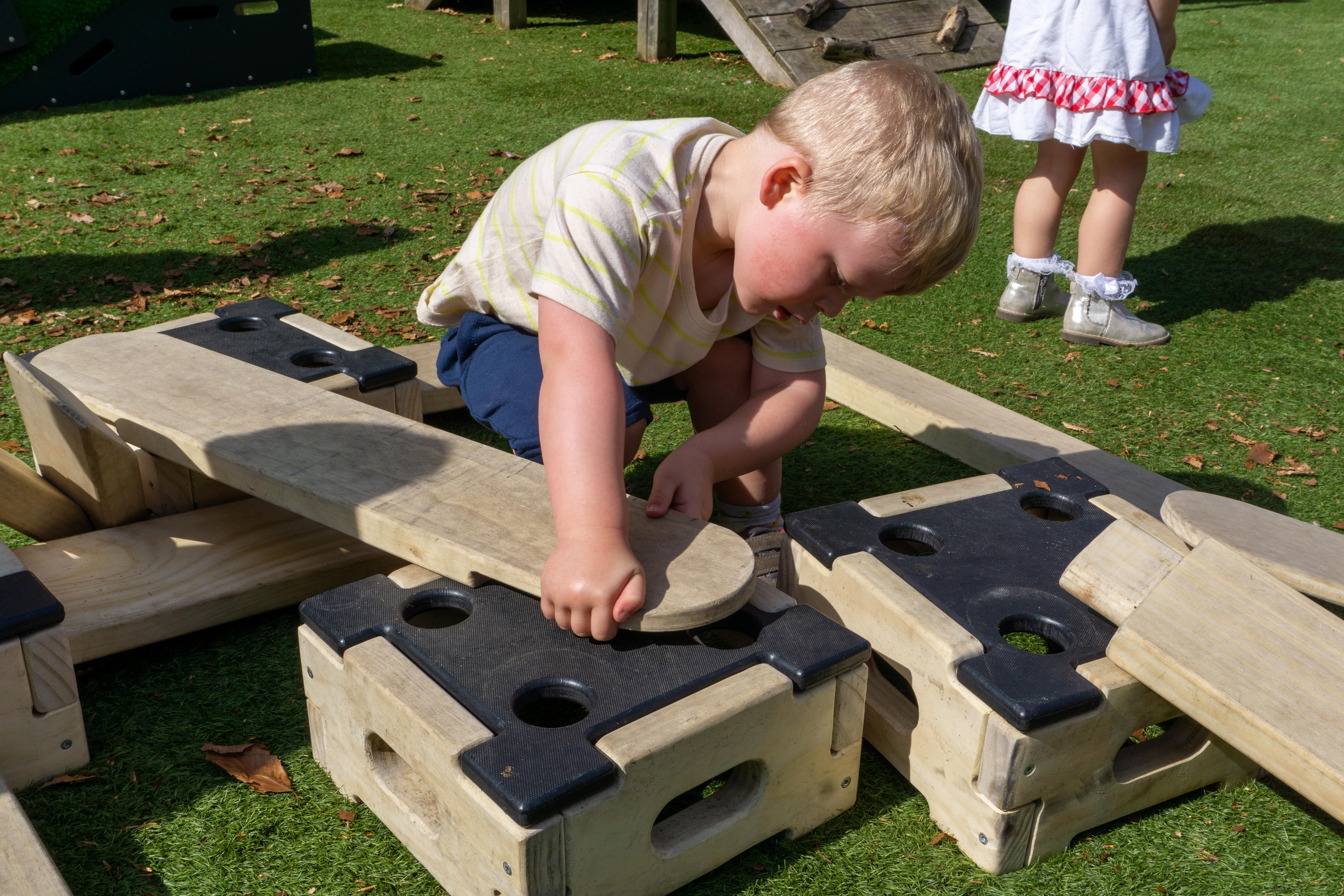 A close up of a little boy connecting a wooden plank to a wooden block. The plank and block are a part of the Play Builder set, which is set up on an artificial grass playground at Kindred White Post, A little girls legs can be seen in the background walking away.