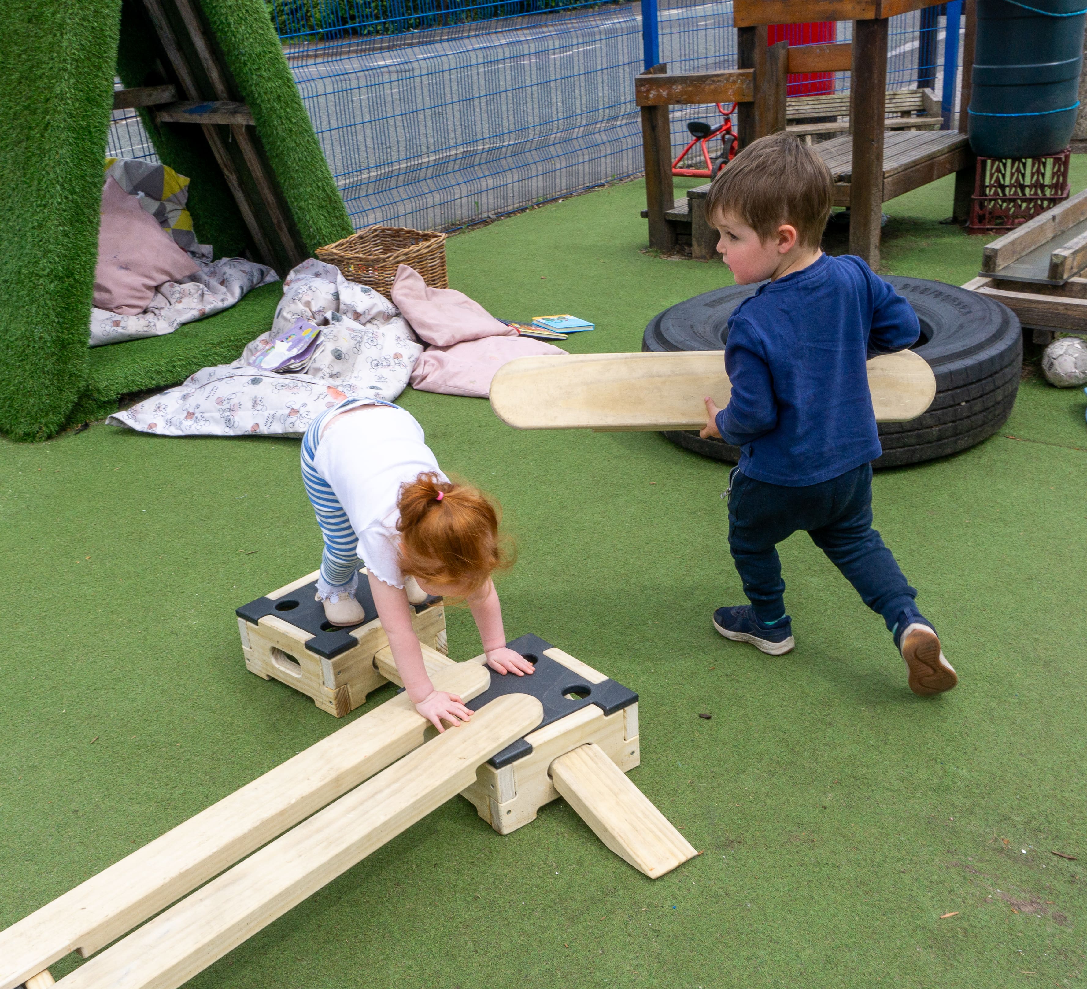 A boy is holding a plank from the Play Builder Apprentice set as he walks around a little girl stood on a Play Builder block. The little girl is bending over as she puts her hands on one block, whilst keeping her feet on another,