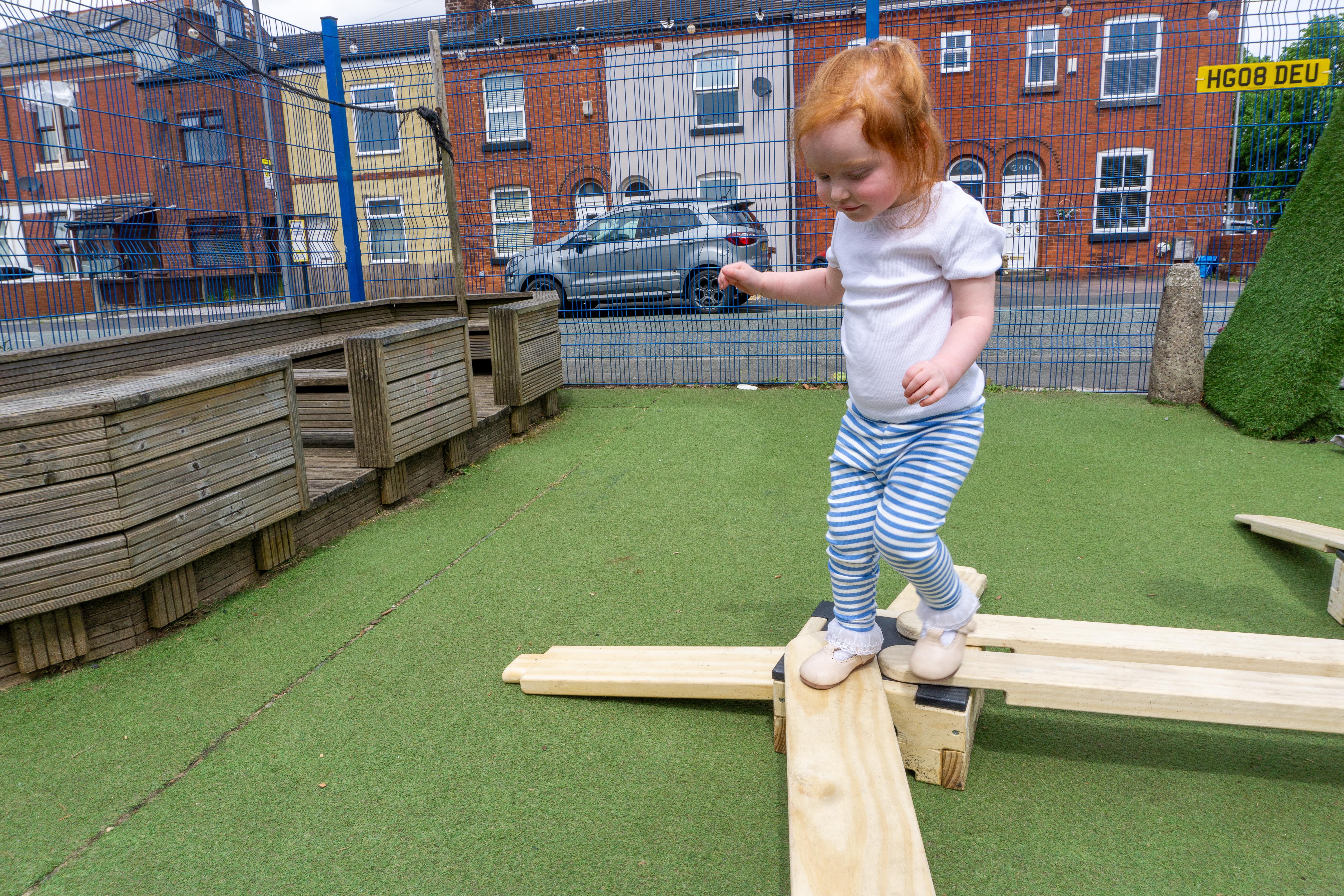 A little girl is walking across wooden planks and she is stood on a wooden block. She is looking down at her feet as she watches where she steps. She has a smile on her face as she engages with the equipment.