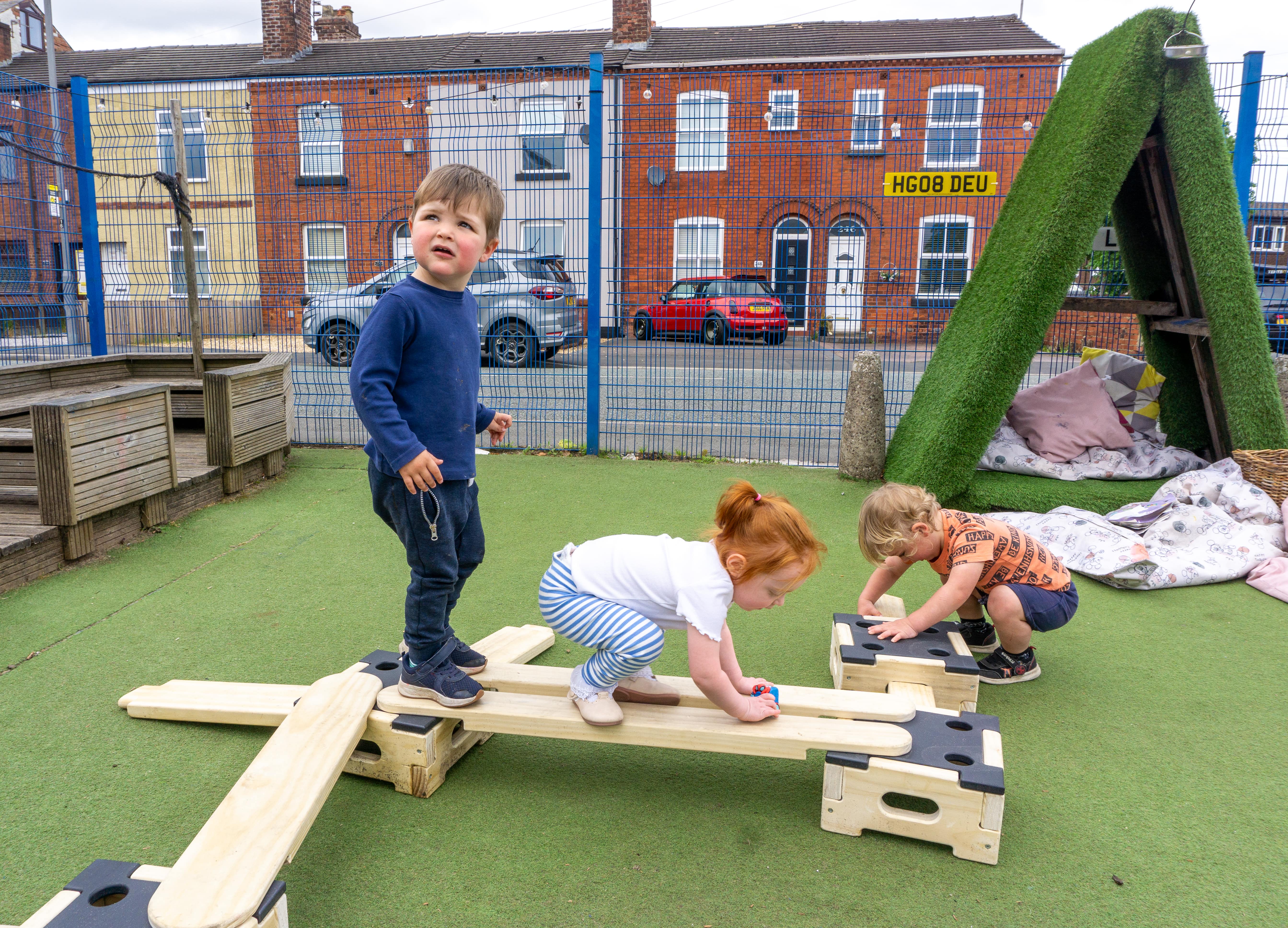 3 children are engaging with the Play Builder Apprentice set. A boy is stood on one of the blocks and is looking towards the camera, whilst a little girl and boy are busy moving the planks and altering it.