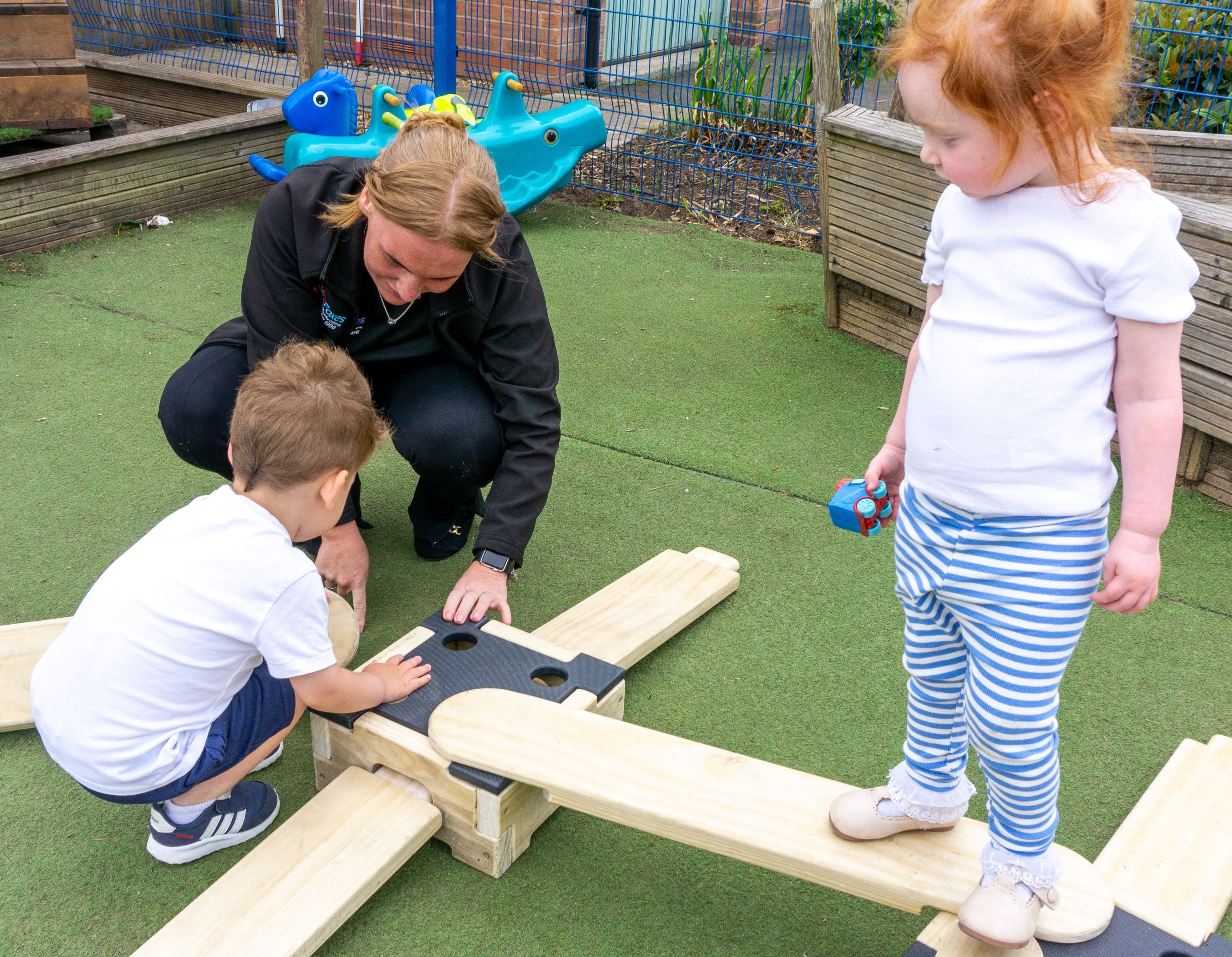 2 children are watching a teacher show them how to connect the wooden planks to the wooden blocks that are a part of the Play Builder Apprentice set. One child is knelt down, getting close to the teacher. The other child is stood on a wooden plank and is watching from above.