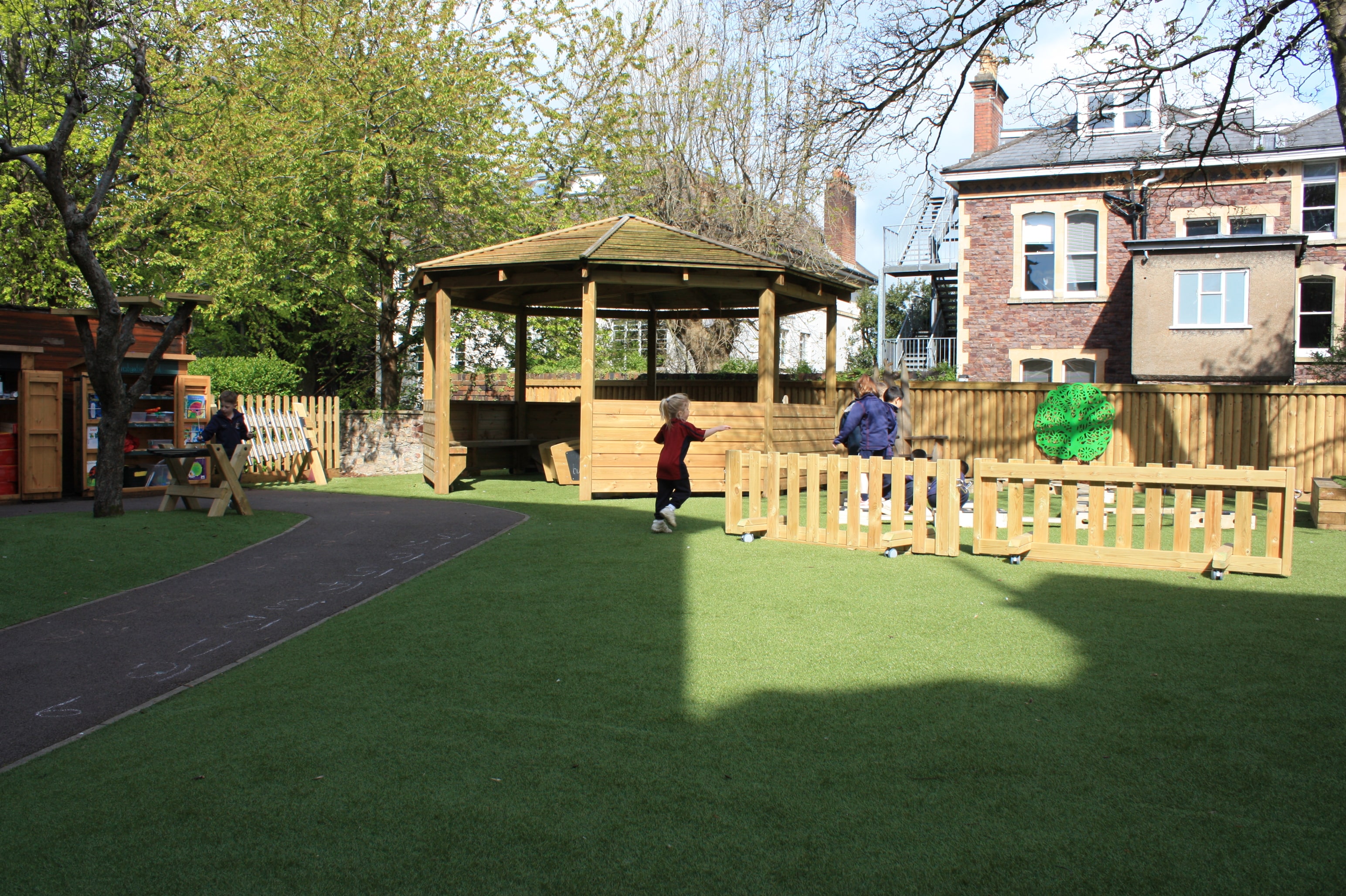 An artificial grass playground with a tarmac path on the left. A wooden canopy can be seen with 6 children to the right playing in a fenced off area. The fences are on wheels, allowing them to be moved,