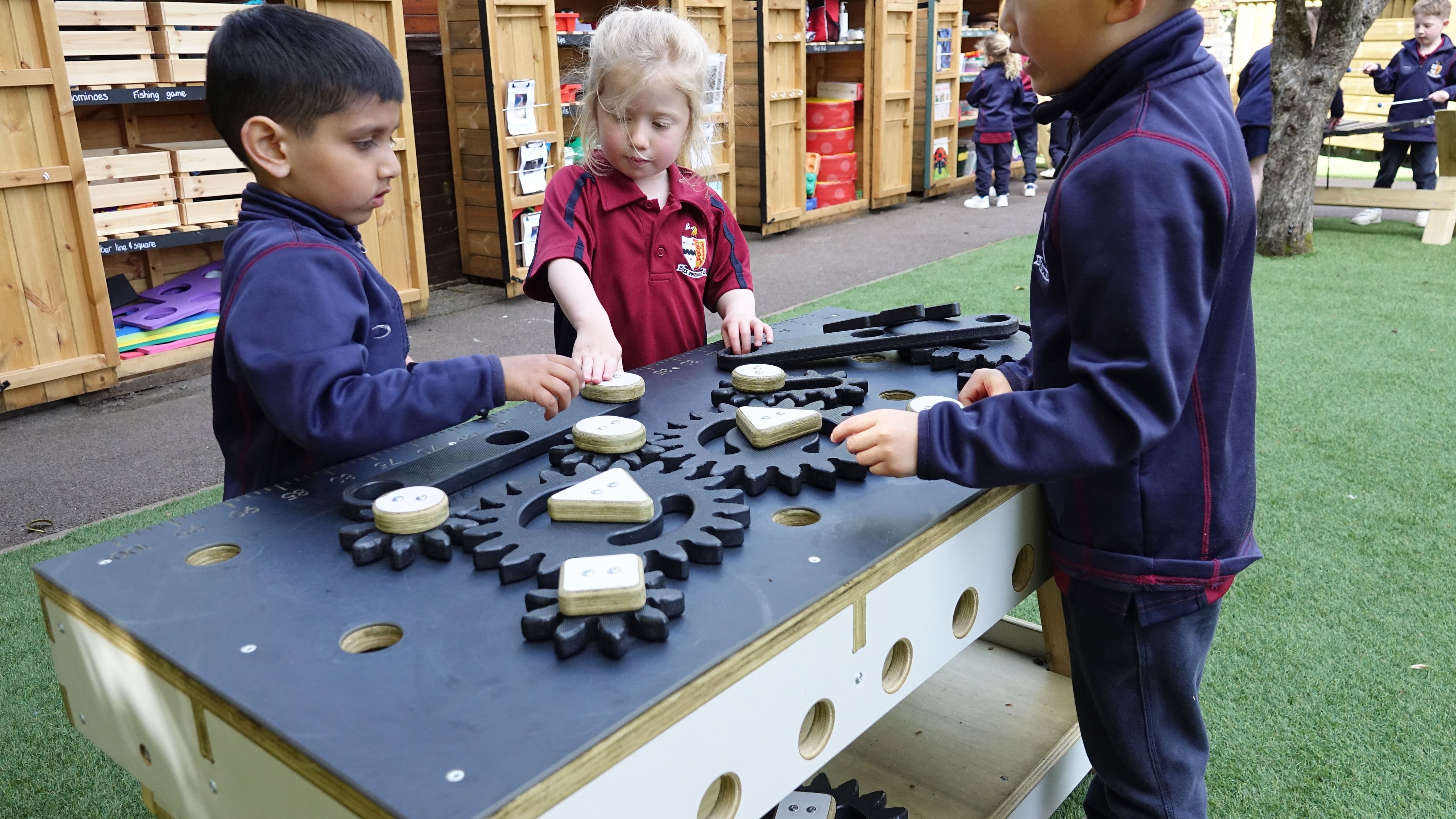 3 children are moving the wooden pieces and cogs on the STEAM table. The top of the table is black, with white sides and a natural wooden shelf.