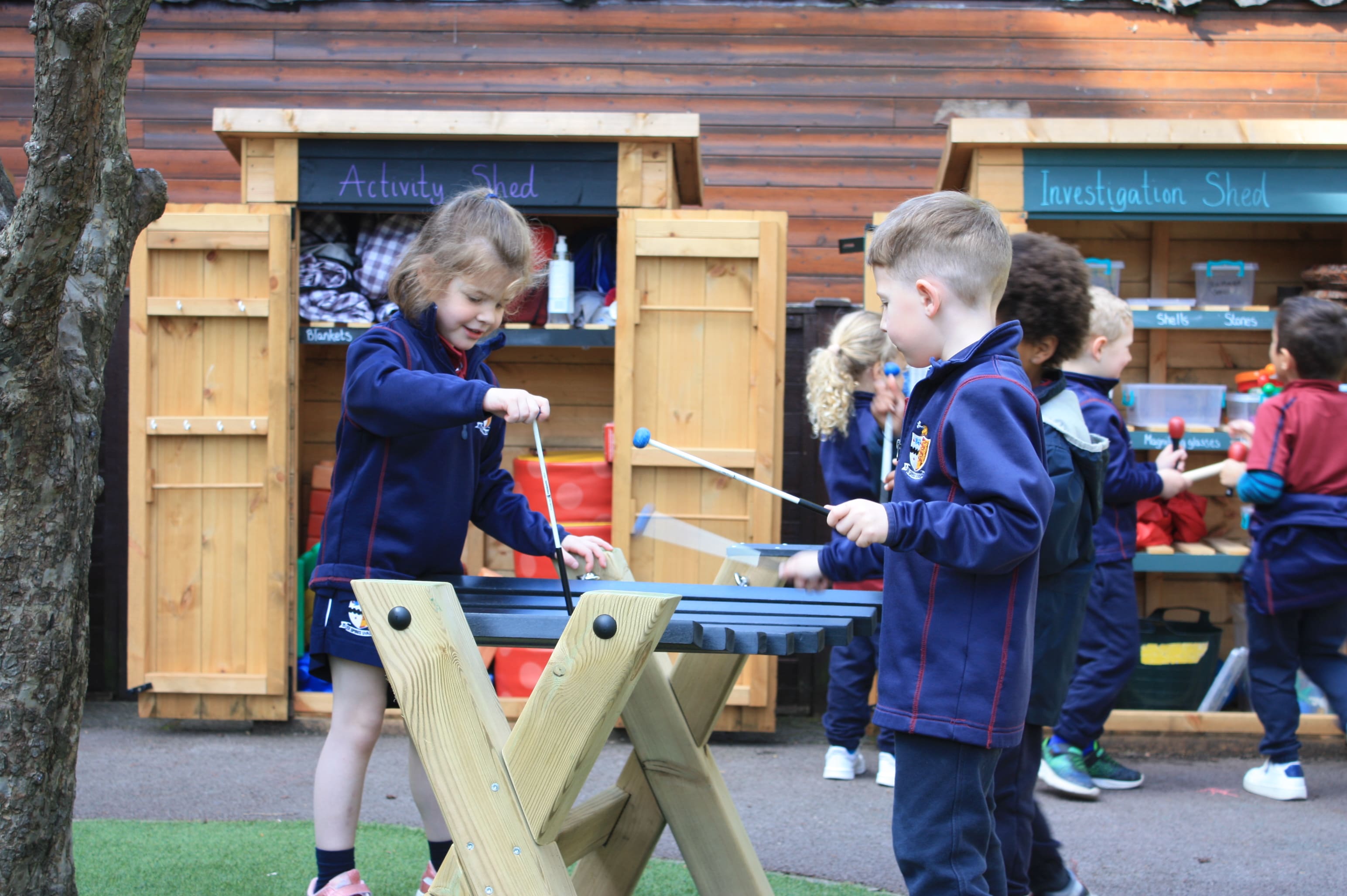 2 children are playing with a mini xylophone and in an area of the playground. Two playground storage cupboards can be seen saying "Activity Shed" and "Investigation Shed" as the two children hit the Xylophone with the xylophone sticks.