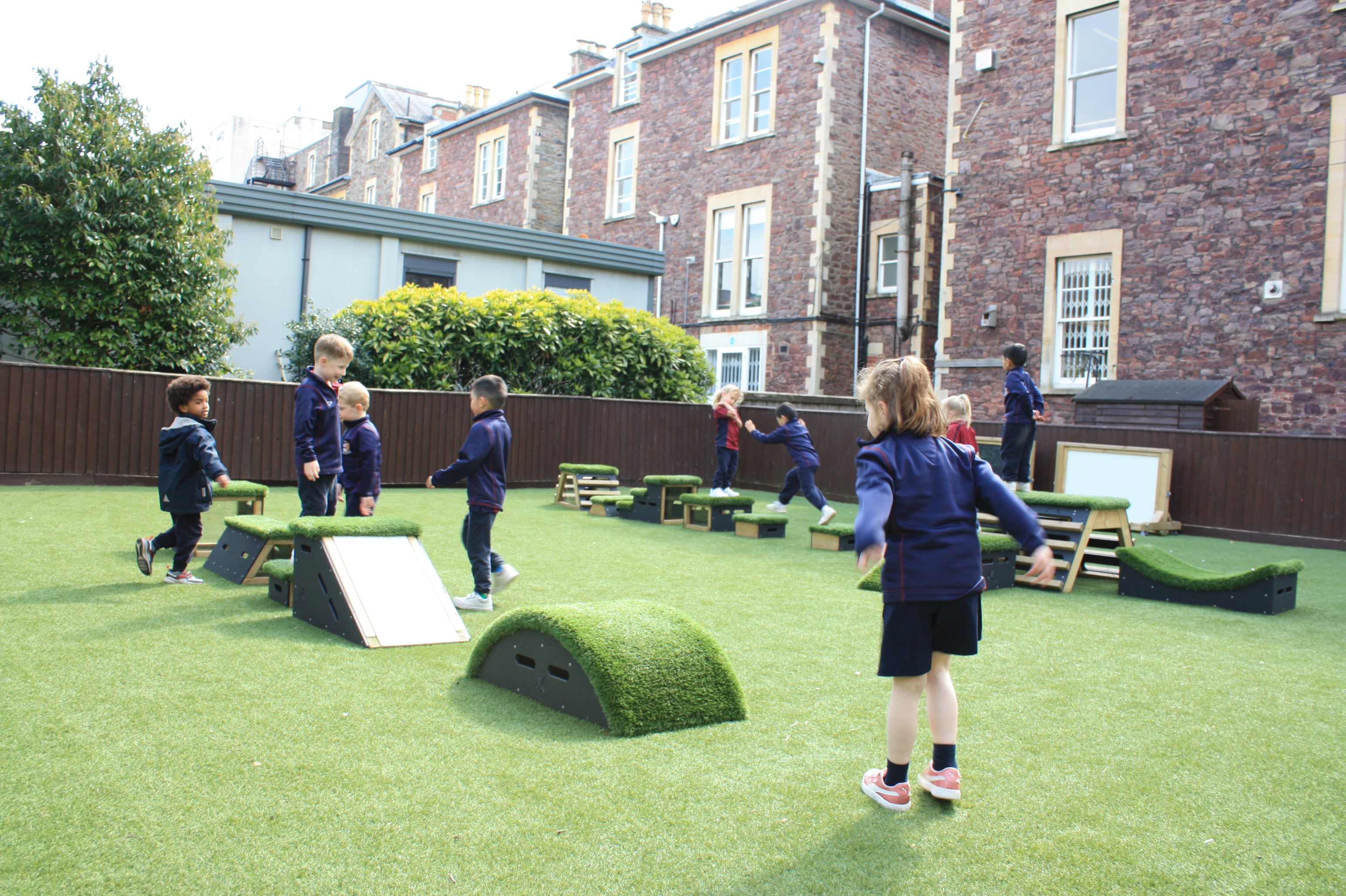A group of children are playing on the Get Set, Go! Blocks. These blocks have wooden sides with an Artificial Grass top and have been set up in 2 straight lines. The children are in two separate groups as they play on them.