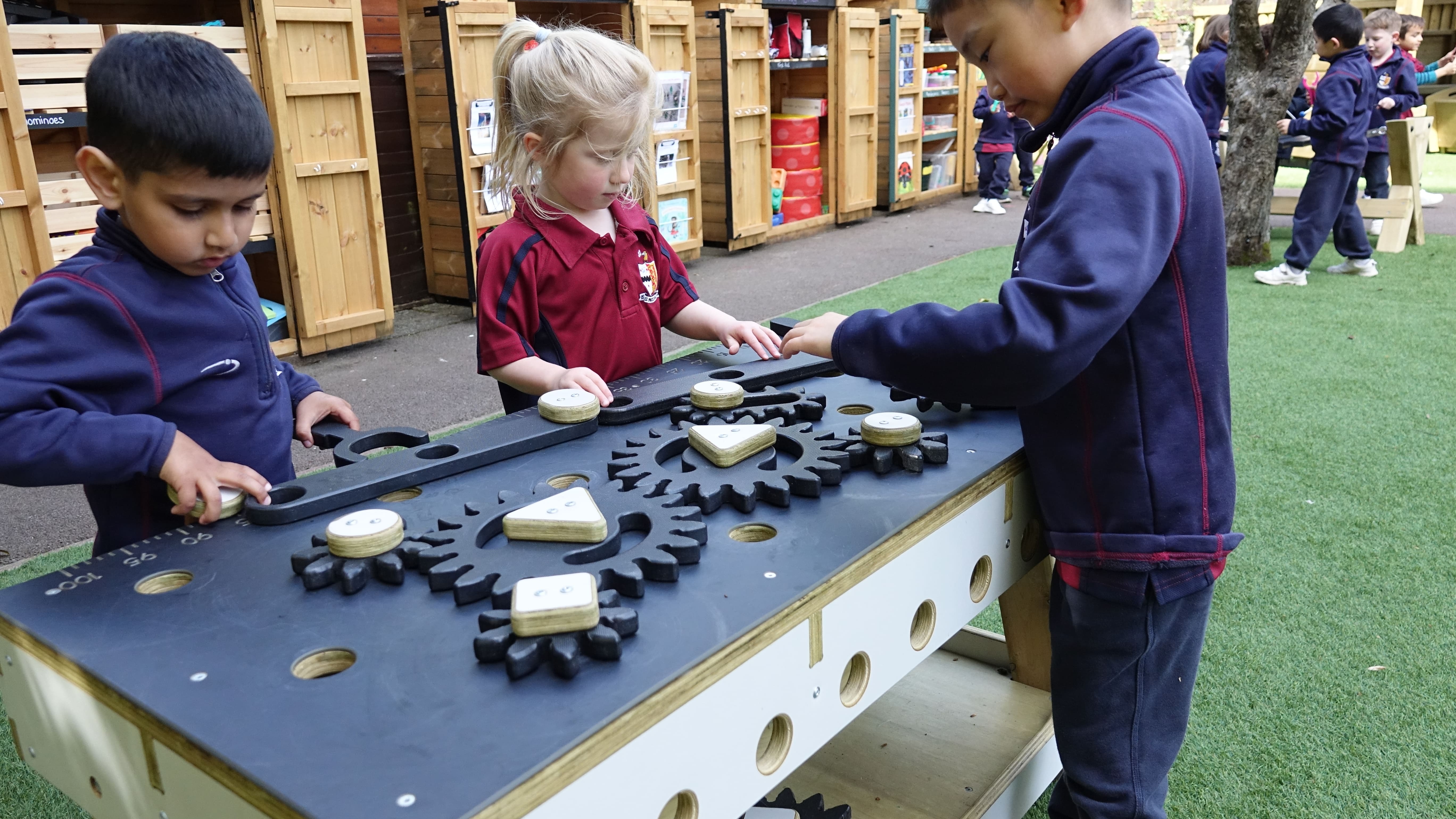 3 children are playing with a wooden table, with a black top and wooden cogs attached to it. The children are moving pieces around on the table to make it flow well.