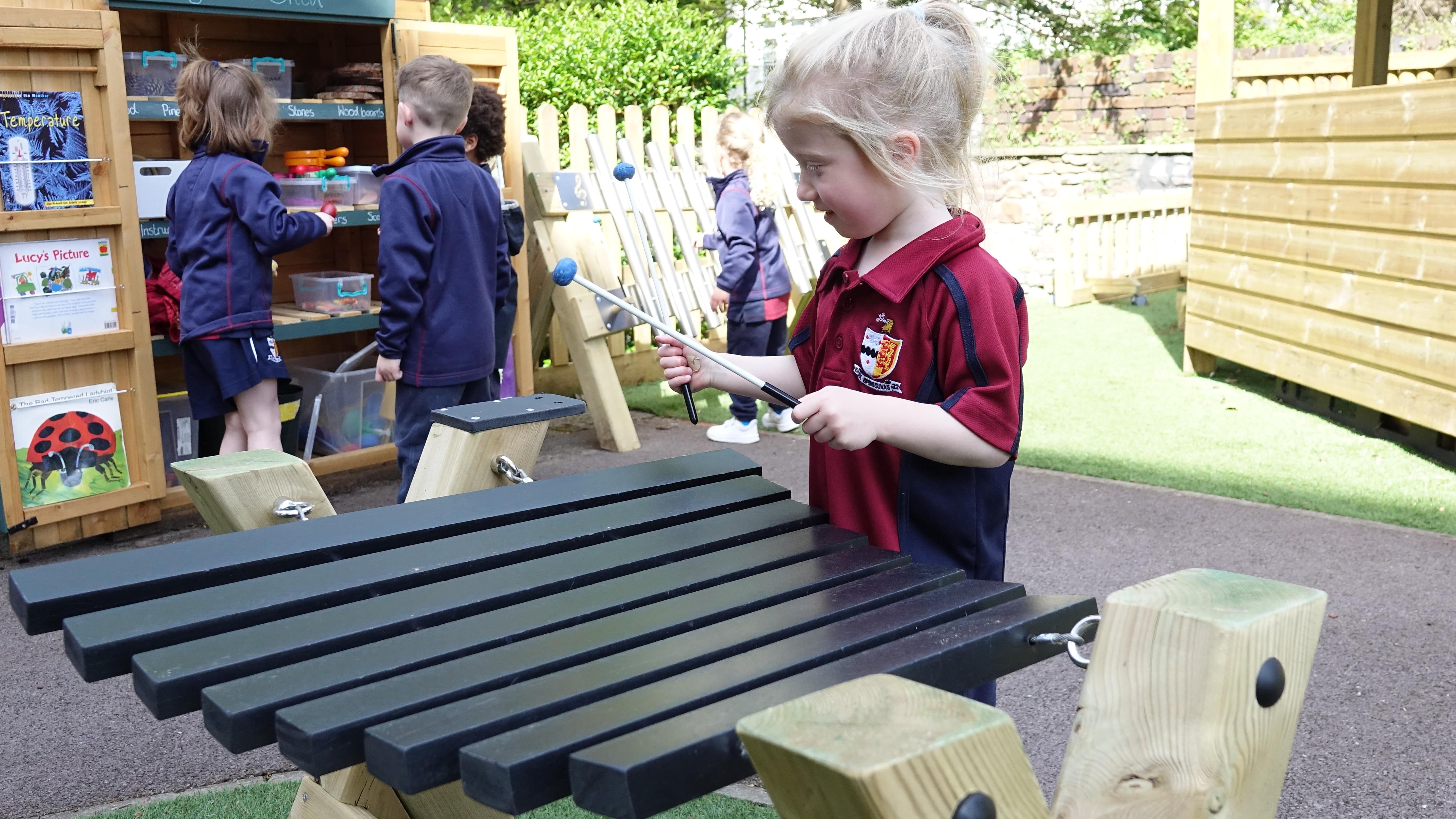 A little girl is playing with a Xylophone, which is on a wooden stand. The girl is holding two sticks to hit each chime as children behind her and looking through a storage cupboard