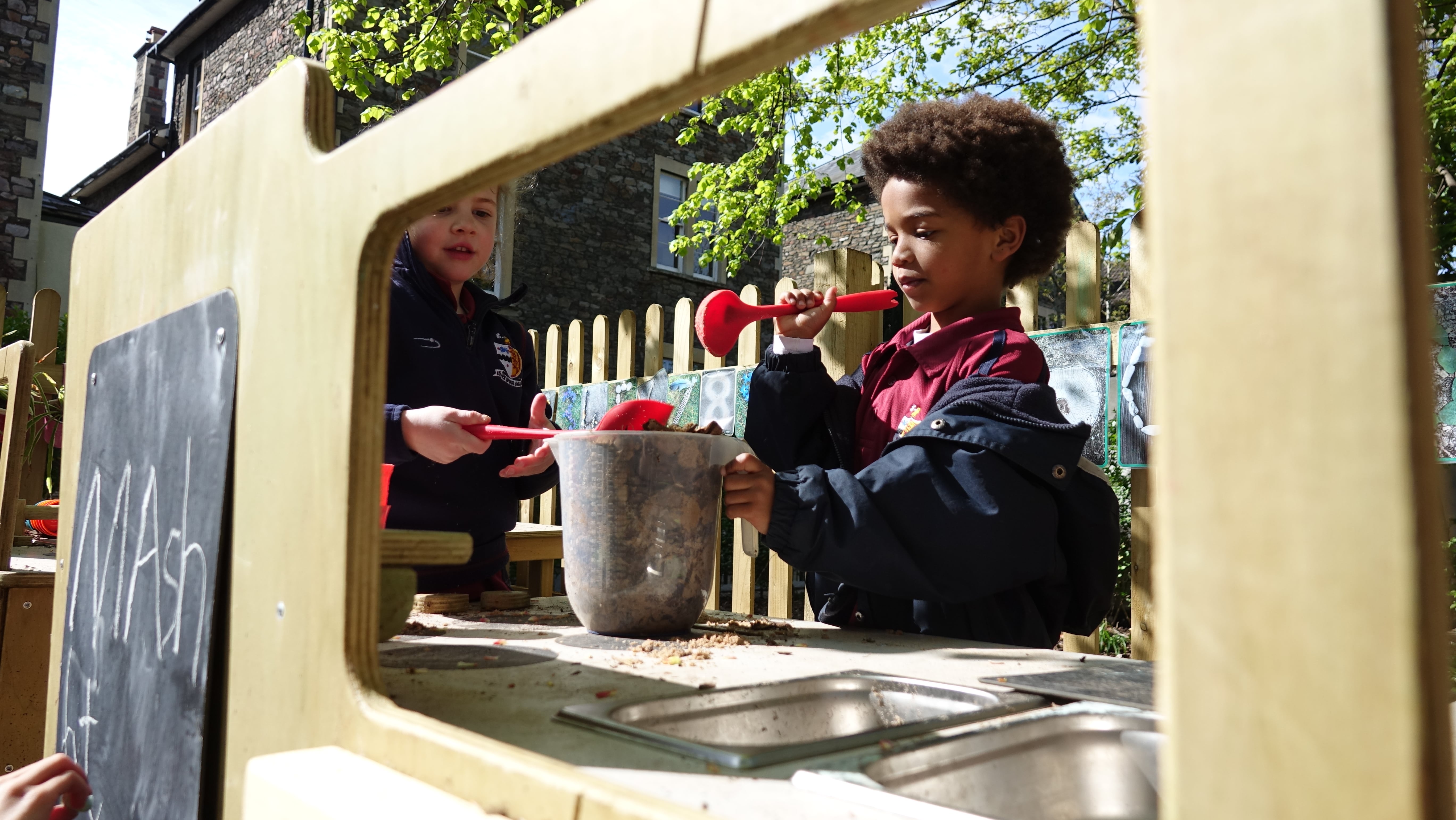 2 Children are using the mud kitchen. The children are filling a plastic jug with leaves and mud and mixing it with a plastic spoon. A chalkboard can be seen on the front, with the word "Mash" written on it,