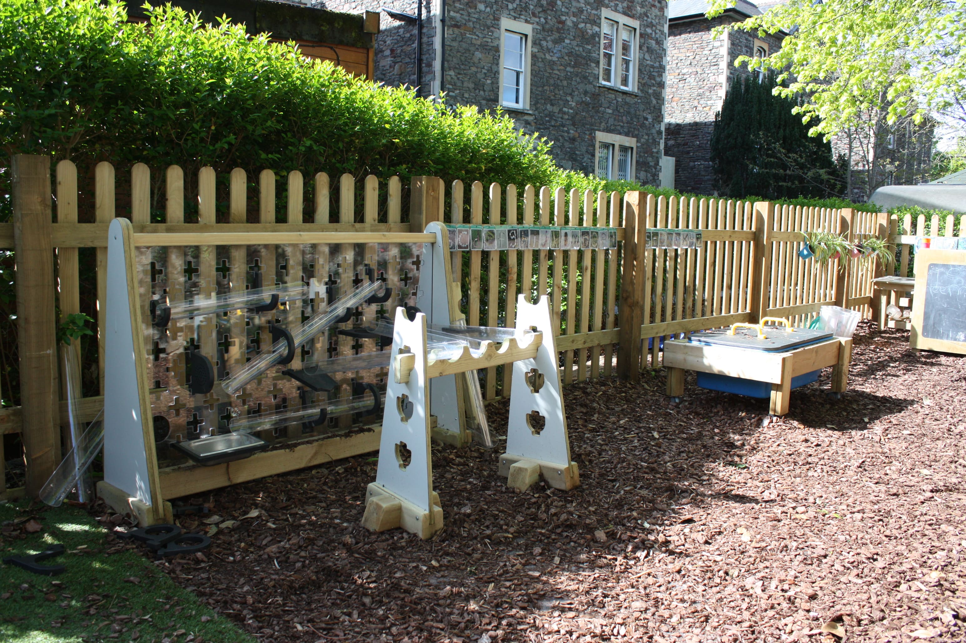 The water wall is shown leaning on a wooden fence, as the plastic tubes can be seen pointing down to a silver tray. Some tubes come out from the wall and lead to another wooden stand. Beside it is the sand table, which has a wooden lid on it and wheels.