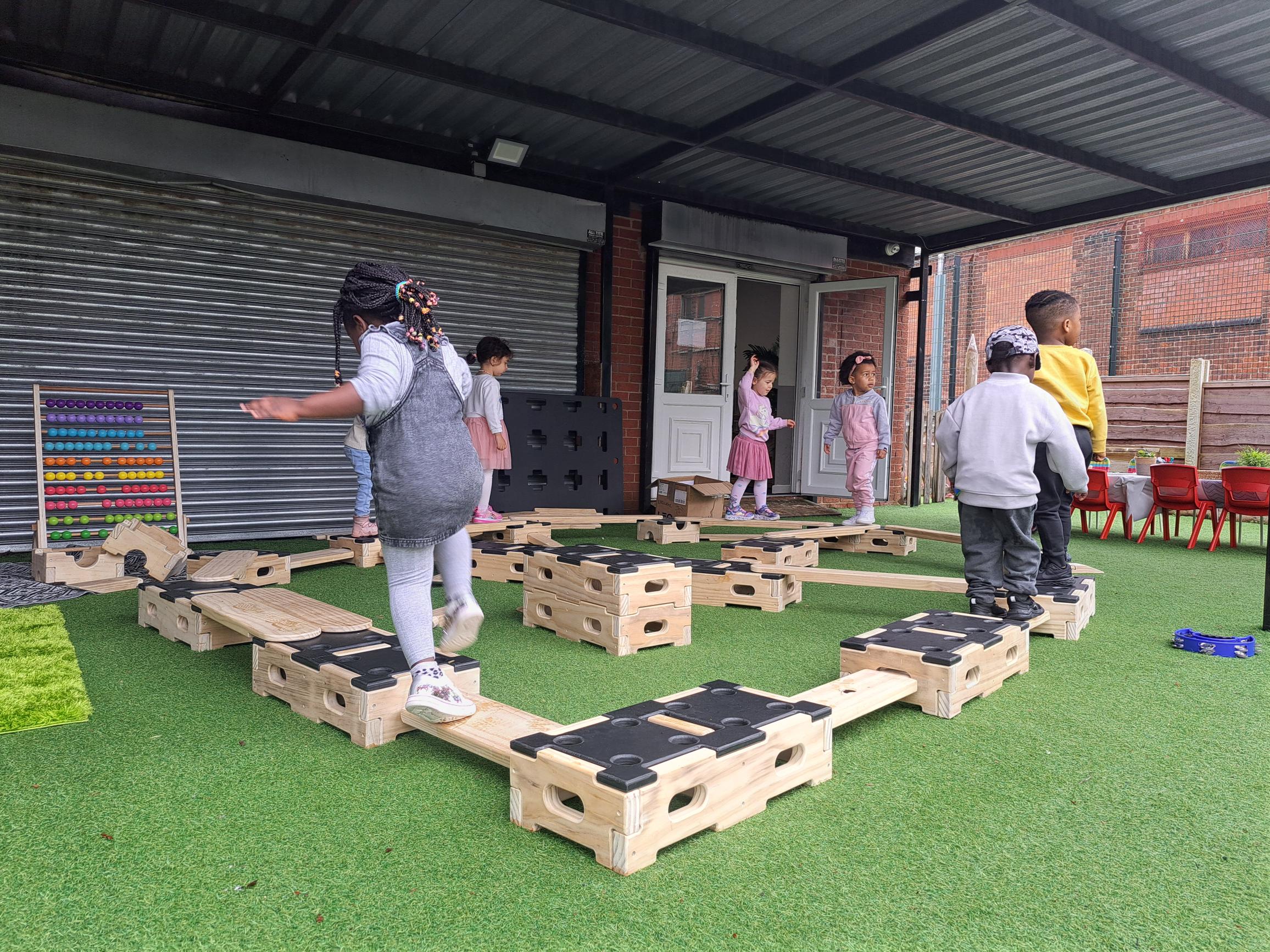 A group of children are balancing and standing on wooden planks and blocks that are included in the Play Builder set. They have created a circuit as the planks and blocks connect to each other.