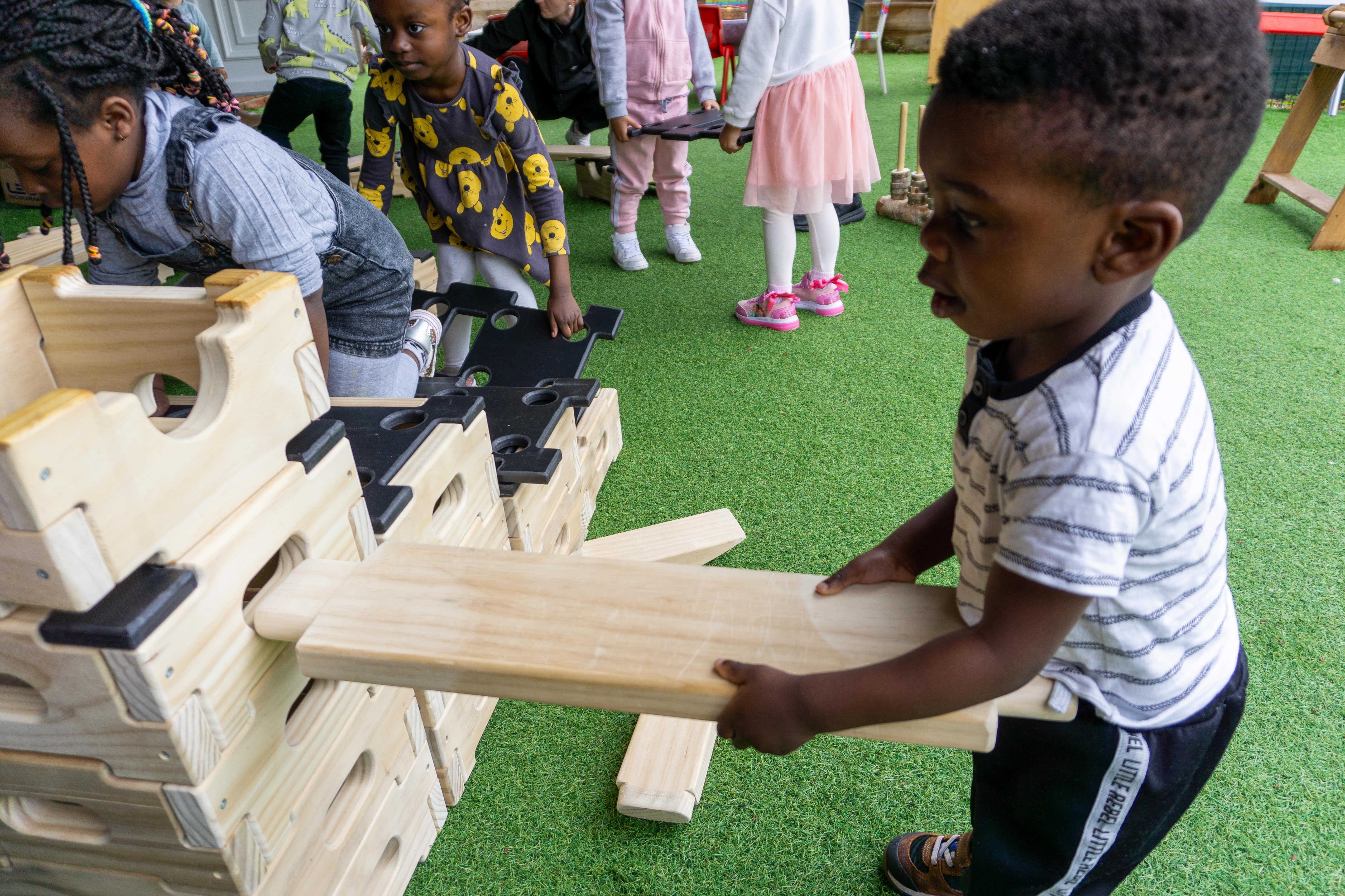 A child is inserting a wooden plank into a wooden block. He is focused on trying to insert the wooden plank perfectly into the whole, allowing it to connect strongly. A group of children behind are watching and collecting other wooden planks and blocks to use.