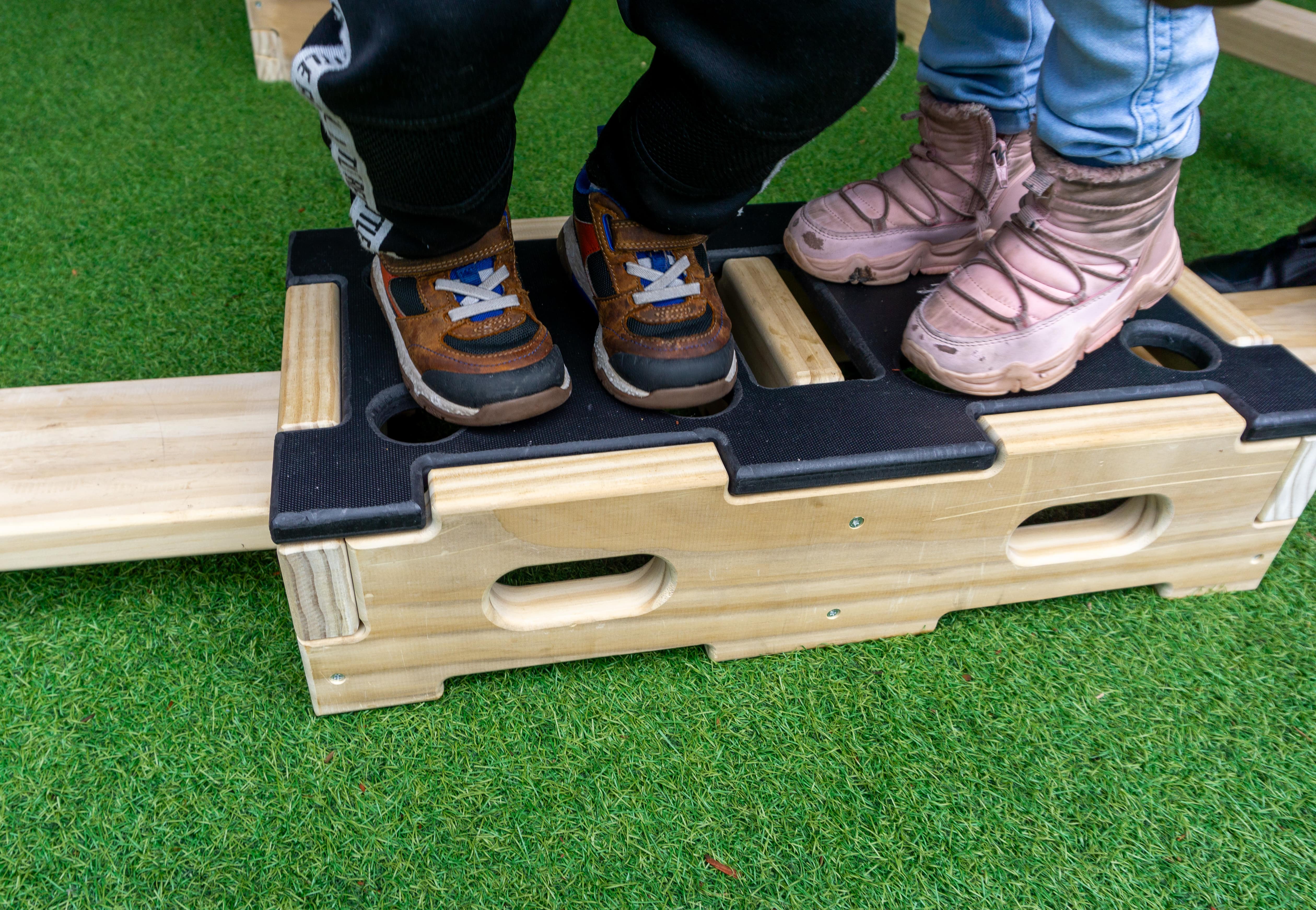 Two children are stood on top of a wooden Play Builder block, which has been placed on artificial grass.