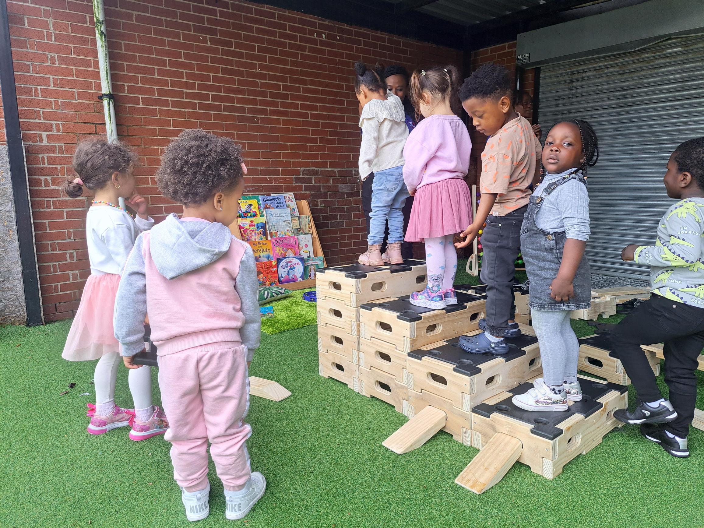 Wooden blocks are stacked on top of each other to create a staircase, with 4 children stood on each step. Each step is one higher than before, creating an ascending order.