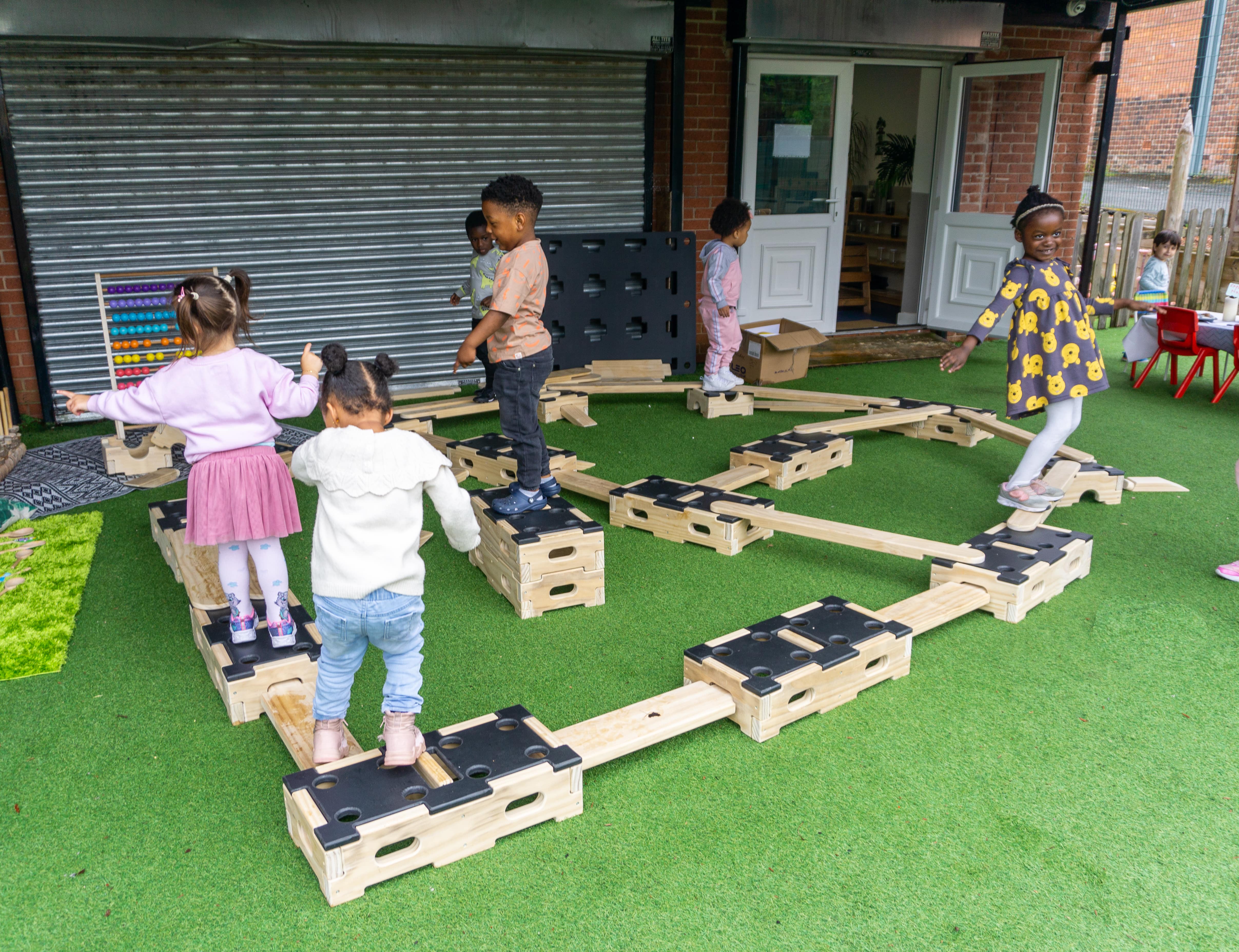 A group of children are completing an obstacle course which has wooden planks connecting to a variety of different wooden blocks. The children are balancing across the planks or standing on a wooden block whilst the surrounding area is covered in artificial grass surfacing.