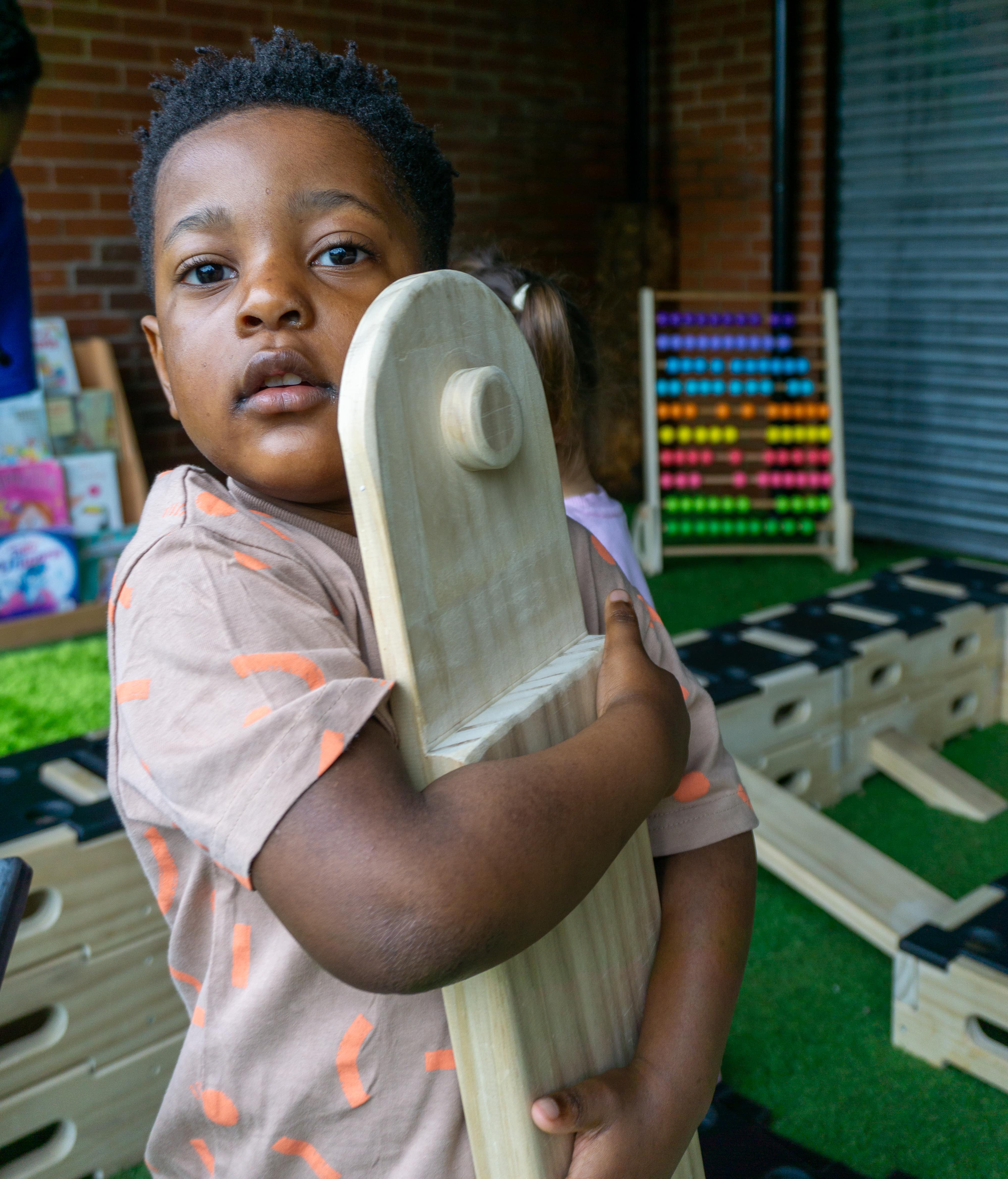 A young boy is holding a wooden plank from the Play Builder set and is looking at the camera. Whilst he's looking at the camera, he's hugging the wooden plank and smiling whilst surrounded by other playground equipment.