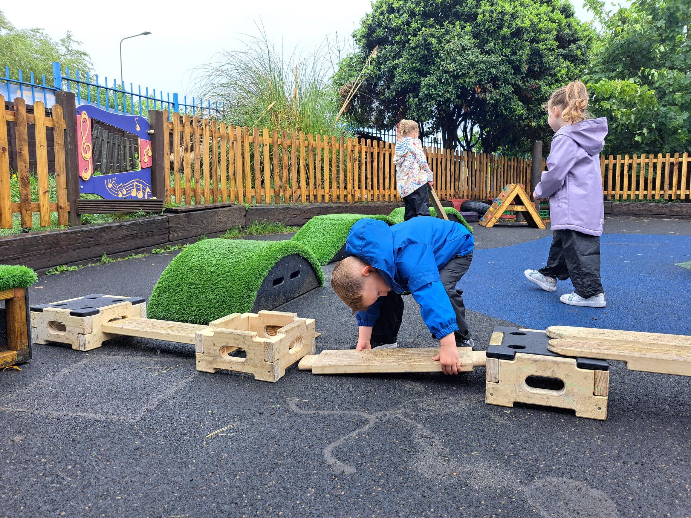 Child connecting a wooden plank to the wooden block contained in the Play Builder sets. Two other children are watching.