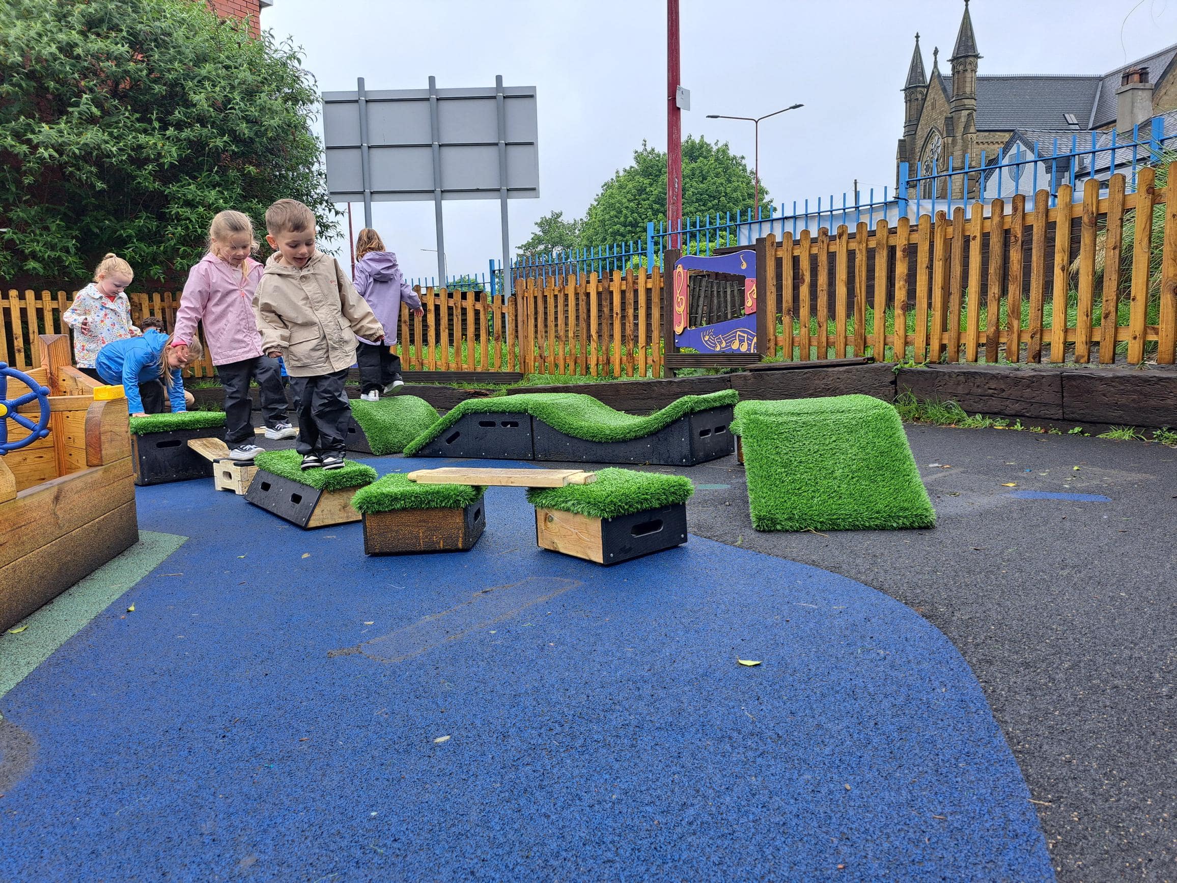 A group of children are picking up wooden planks and blocks as they move them around a playground to create an obstacle course. The children look excited and happy as they build.