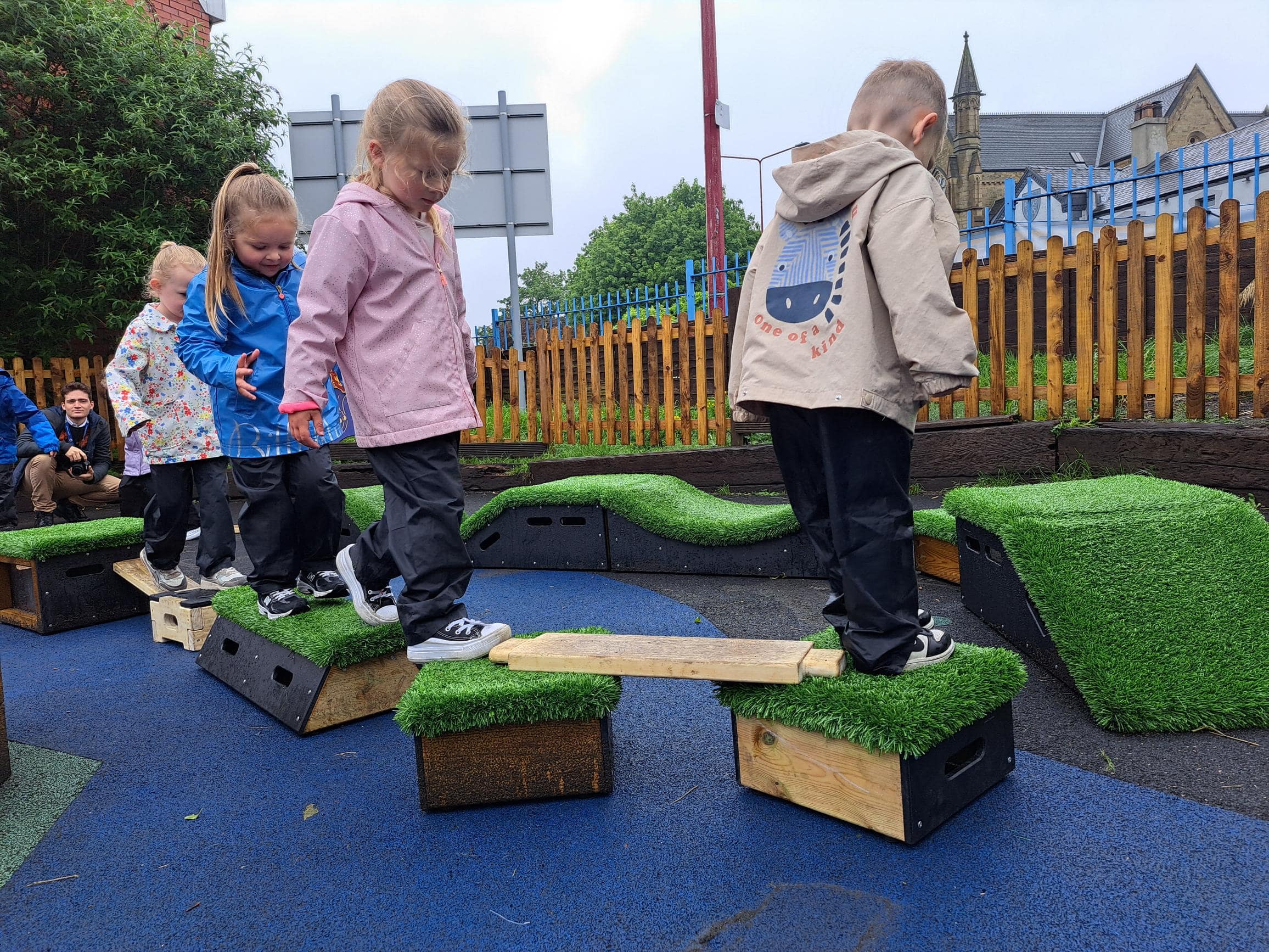 A group of children are completing an obstacle course made from wooden planks and wooden blocks. Some of the wooden blocks are black with artificial grass laid on top of them. The children are in a single file line.