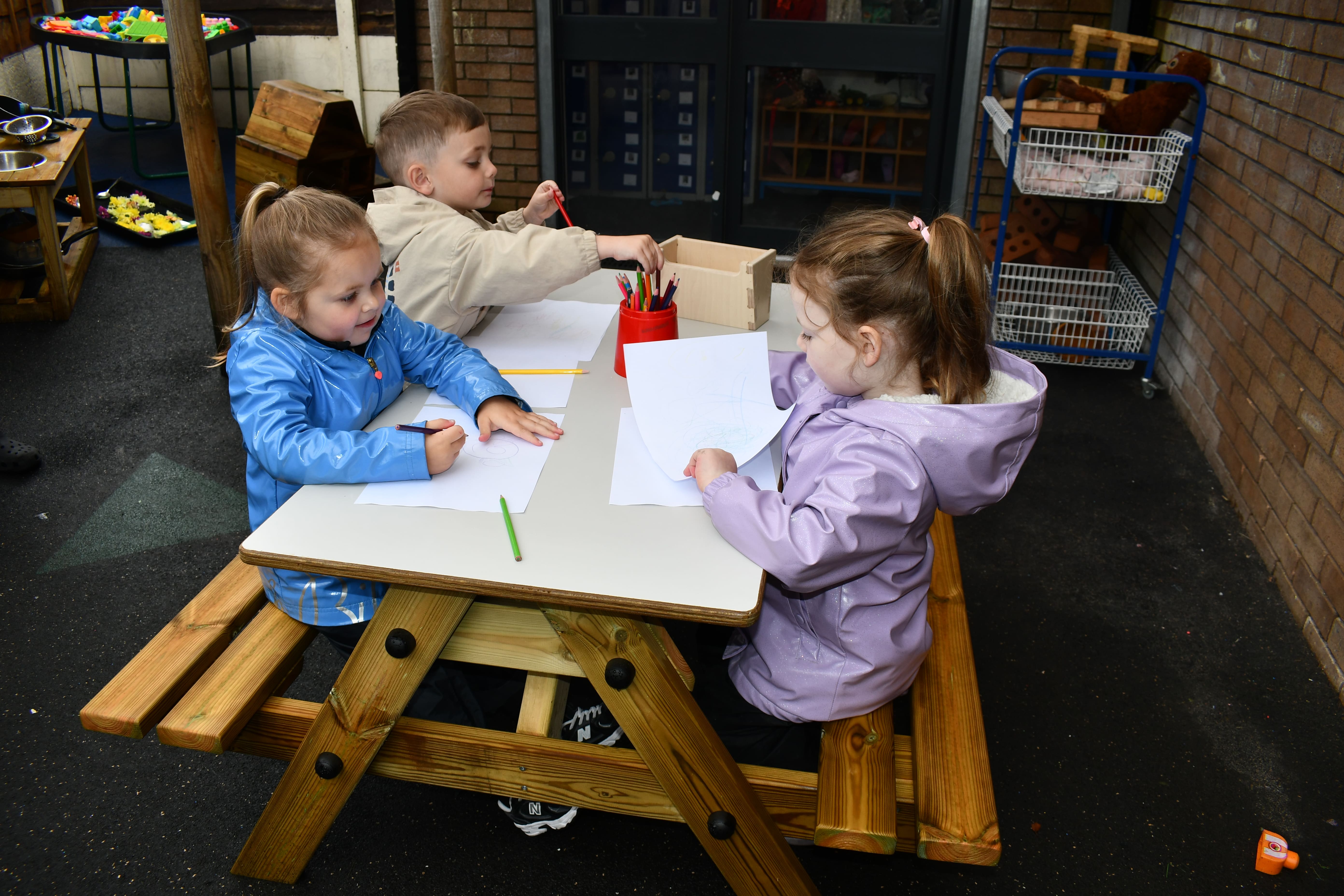3 children are drawing and colouring in on the EYFS Picnic Bench. The bench is placed on top of a hard surface. The top of the table is covered in paper and pencils.
