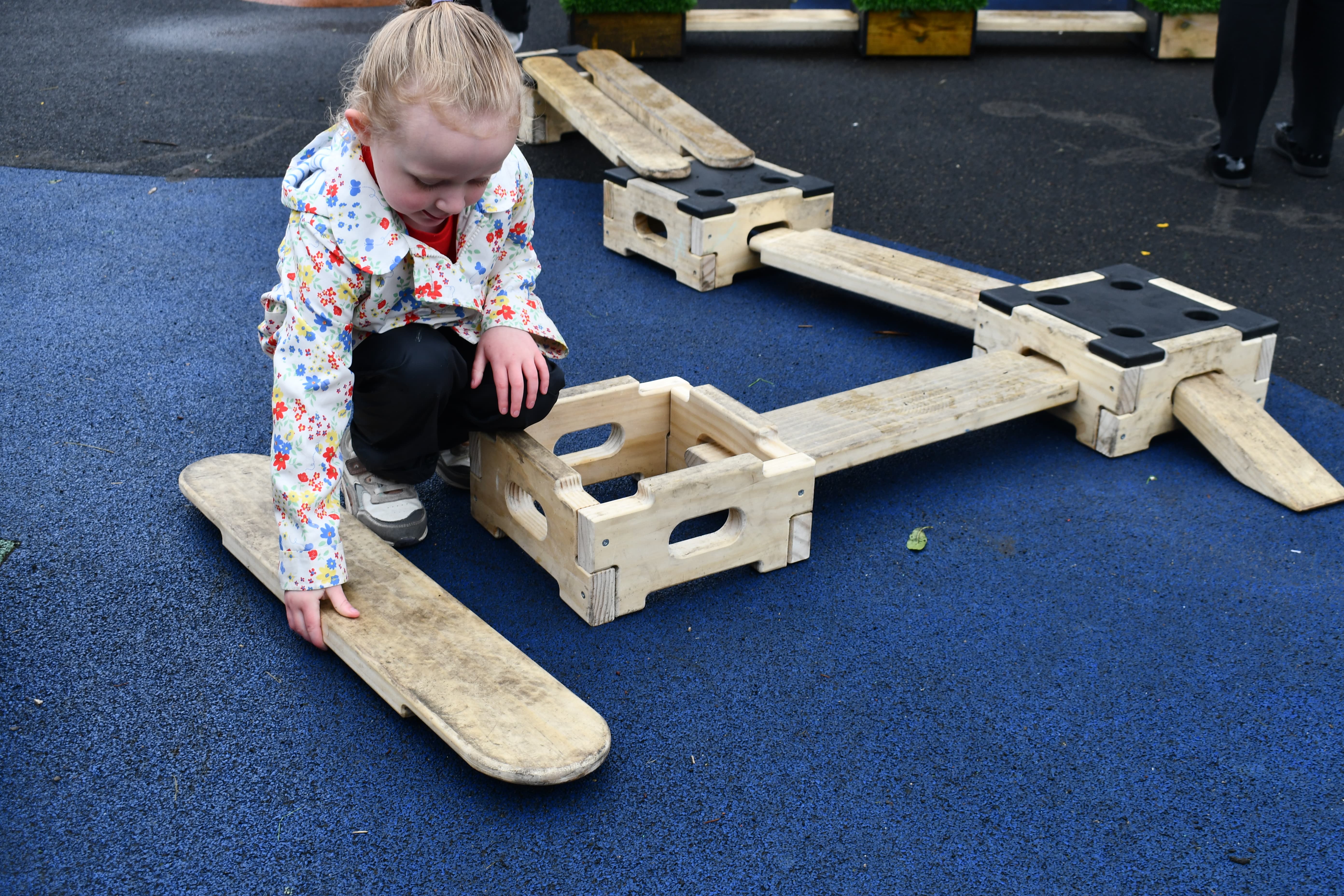 A little girl is playing with wooden planks and boxes from the Play Builder set. She is kneeling down and placing one hand on a plank and the other on her knee. The Play Builder set is laid out on a wetpour surface.