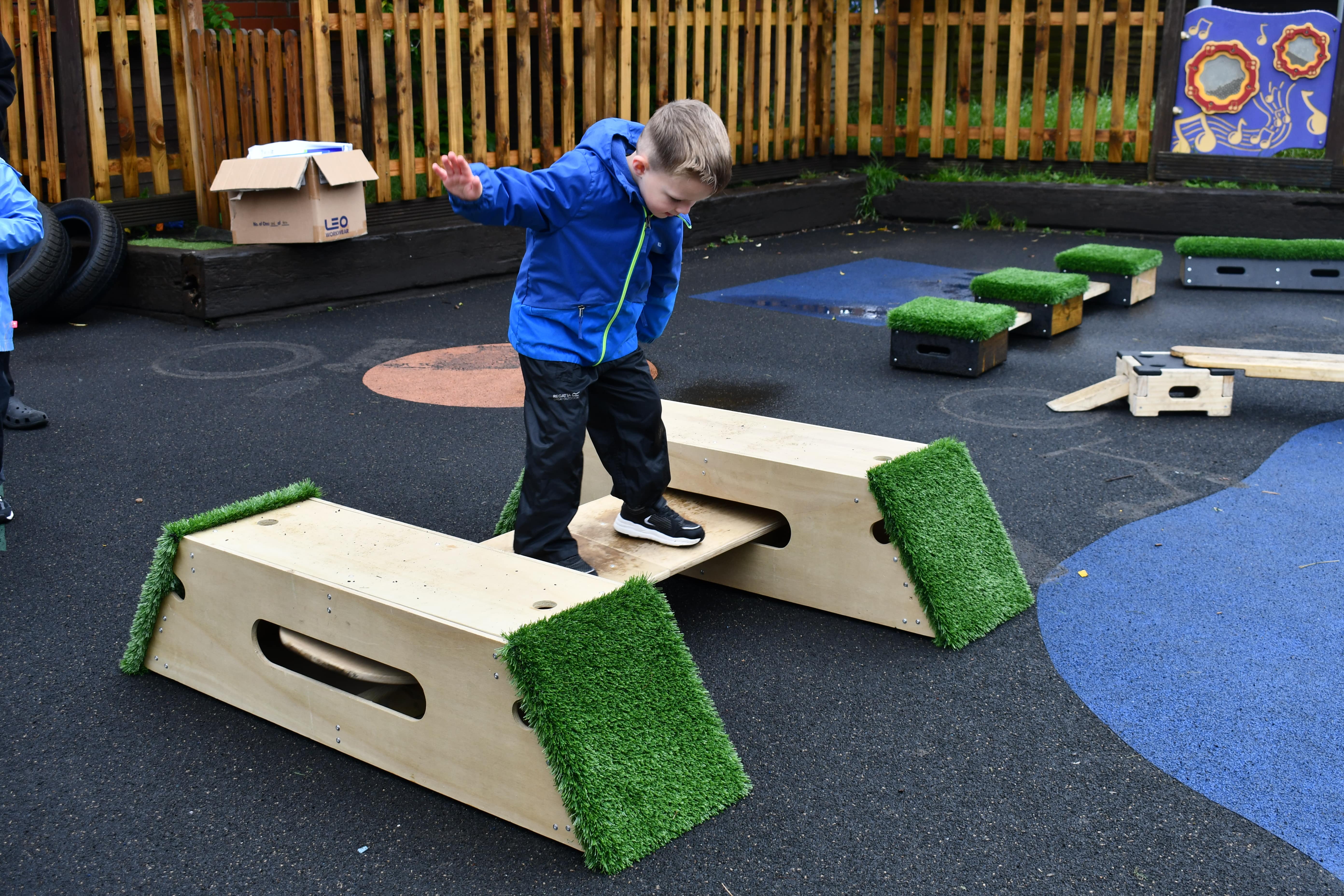 A child is trying to balance on a board which is connected to two wooden blocks, creating a platform for rocking on. The child is sticking his arms out to try and balance whilst looking down at his feet.