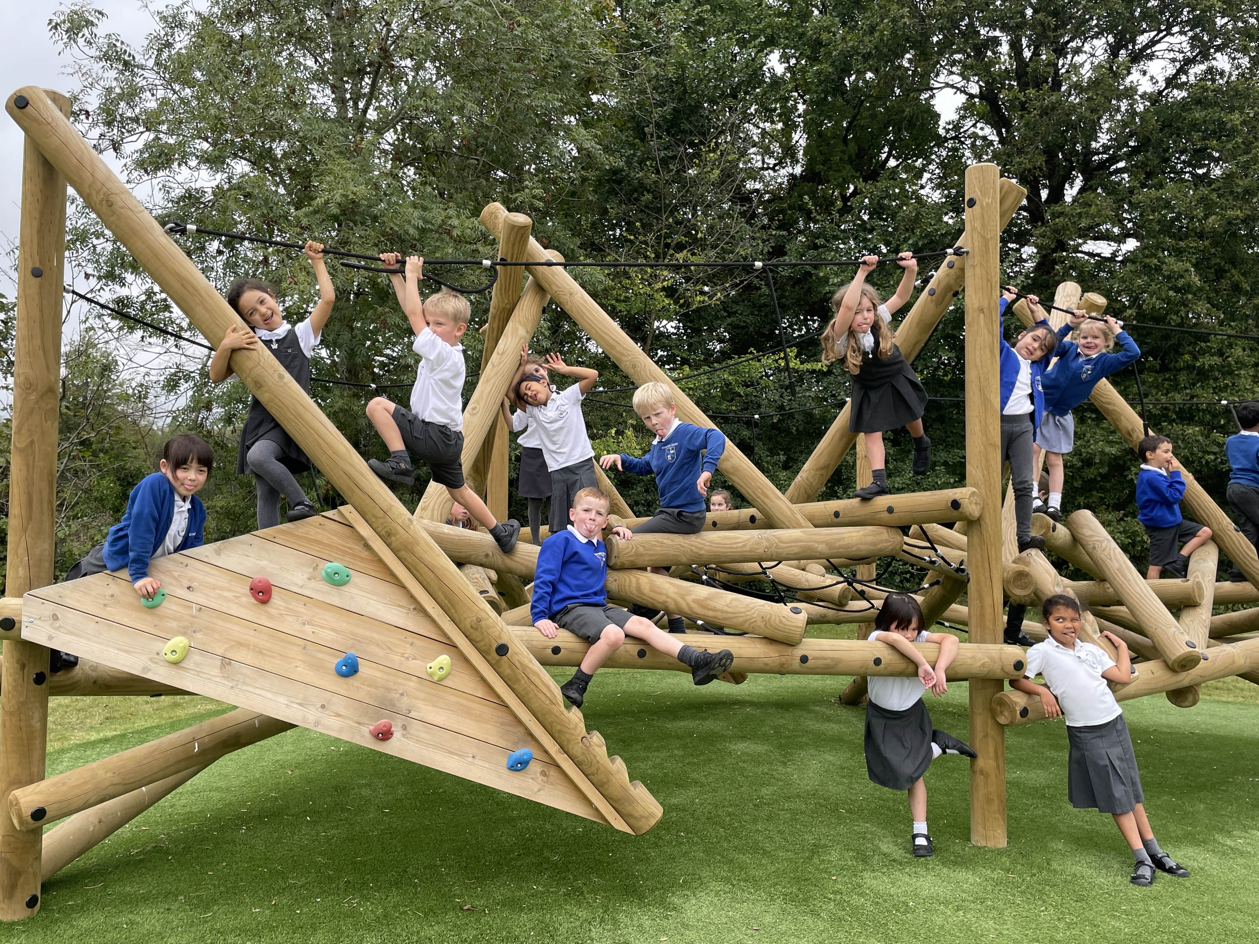 A photo showing a bunch of kids playing on a wooden climbing frame whilst pulling funny faces at the camera. The equipment has been installed on top of artificial grass.