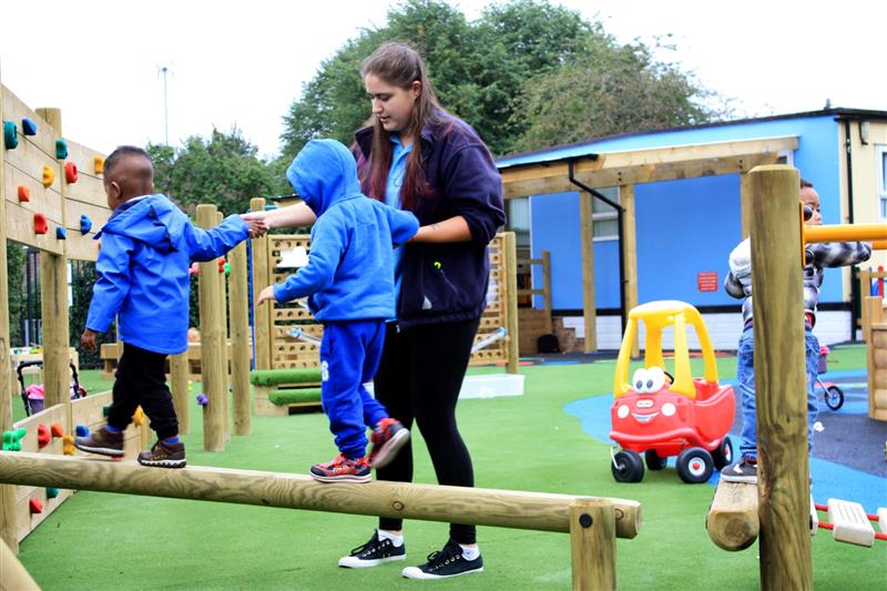 a teacher helps two nursery aged pupils cross the timber balance beam whilst they both wear blue waterproof coats
