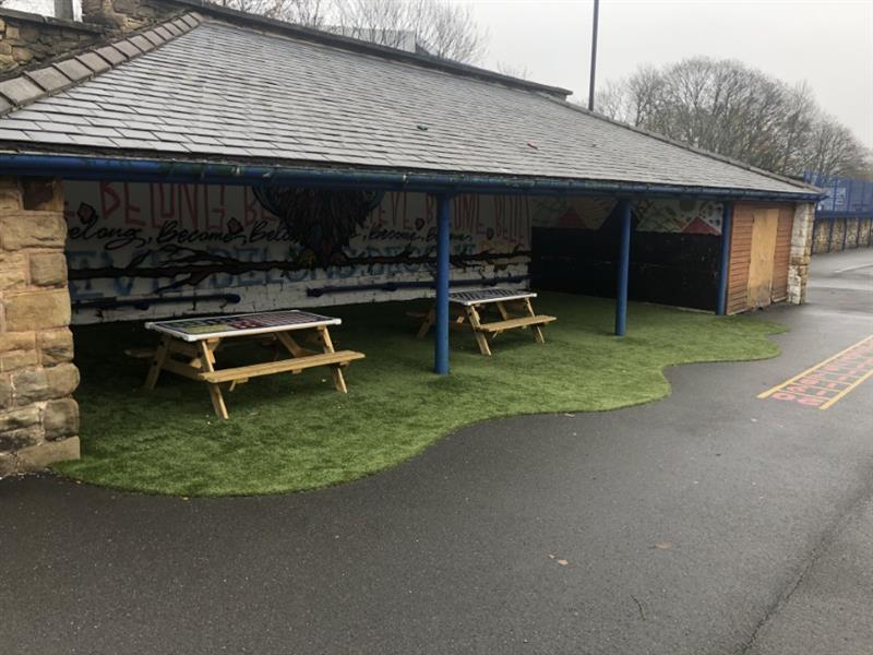 Artificial grass and 2 picnic benches installed under a blue steel canopy with a graffitied brick wall at the rear